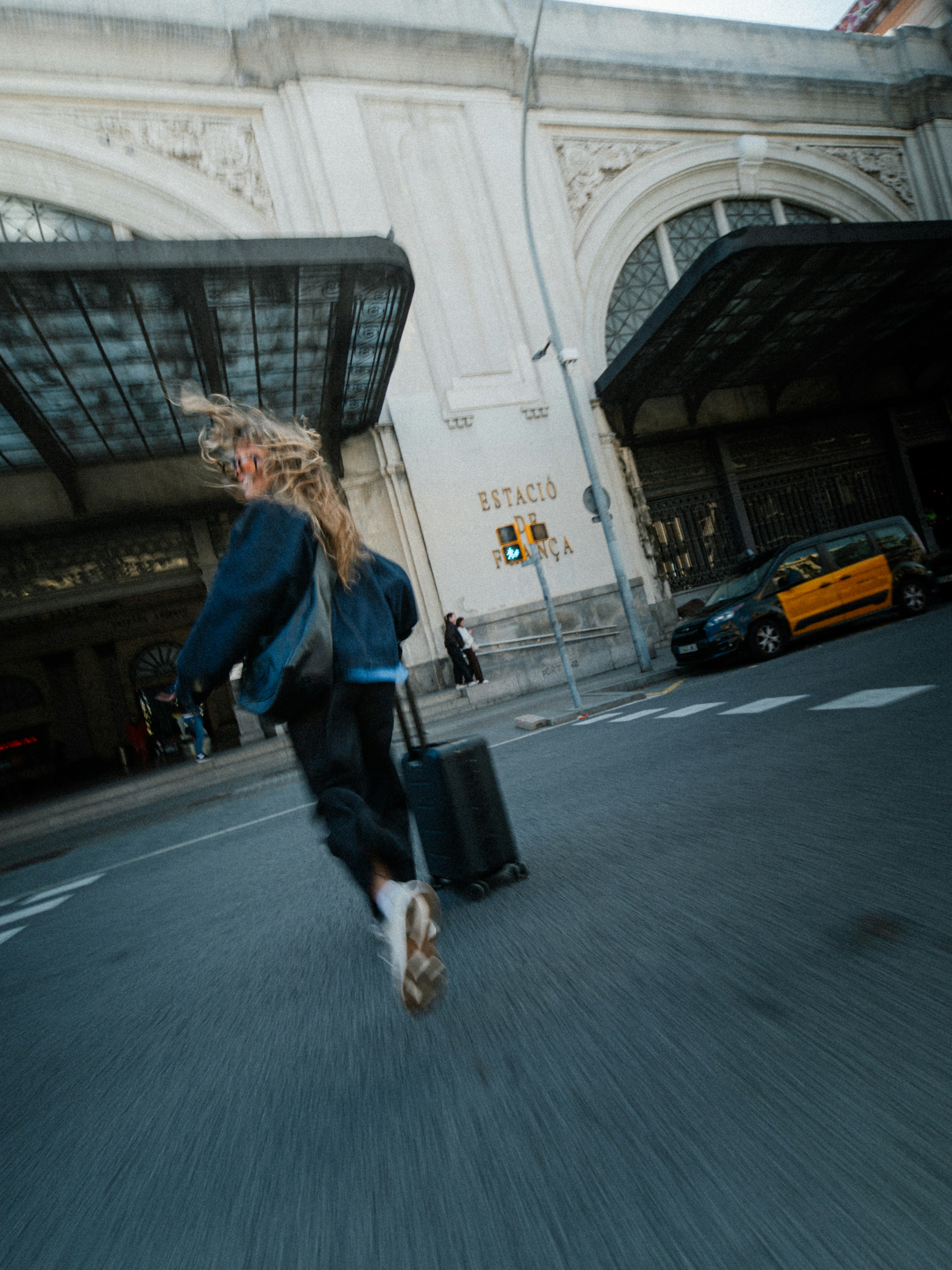 Una persona con una maleta camina hacia un edificio grande y ornamentado con ventanas en arco y un taxi aparcado cerca.