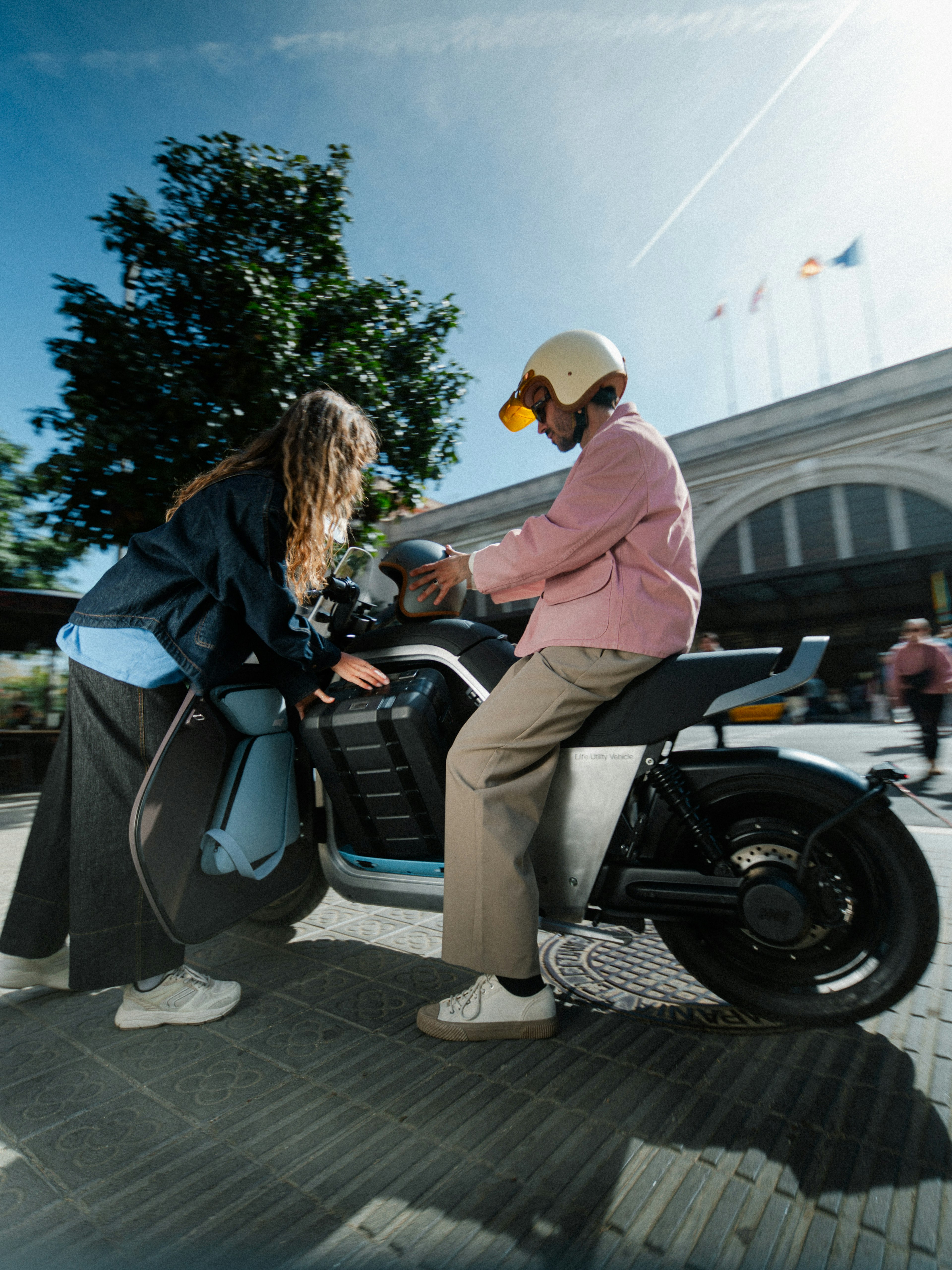 Un hombre con casco en una motocicleta hablando con una mujer frente a un edificio, con banderas y árboles al fondo.
