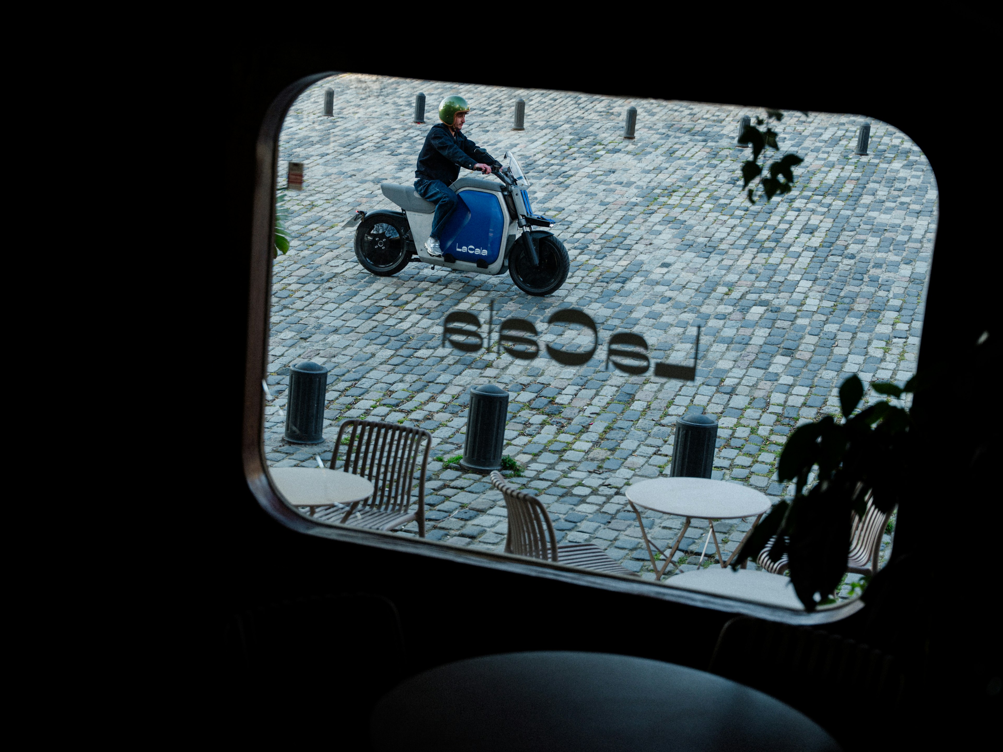 Vista posteriore di una Vespa blu appoggiata a un muro di pietra, circondata da sedie e tavoli in un caffè all'aperto.