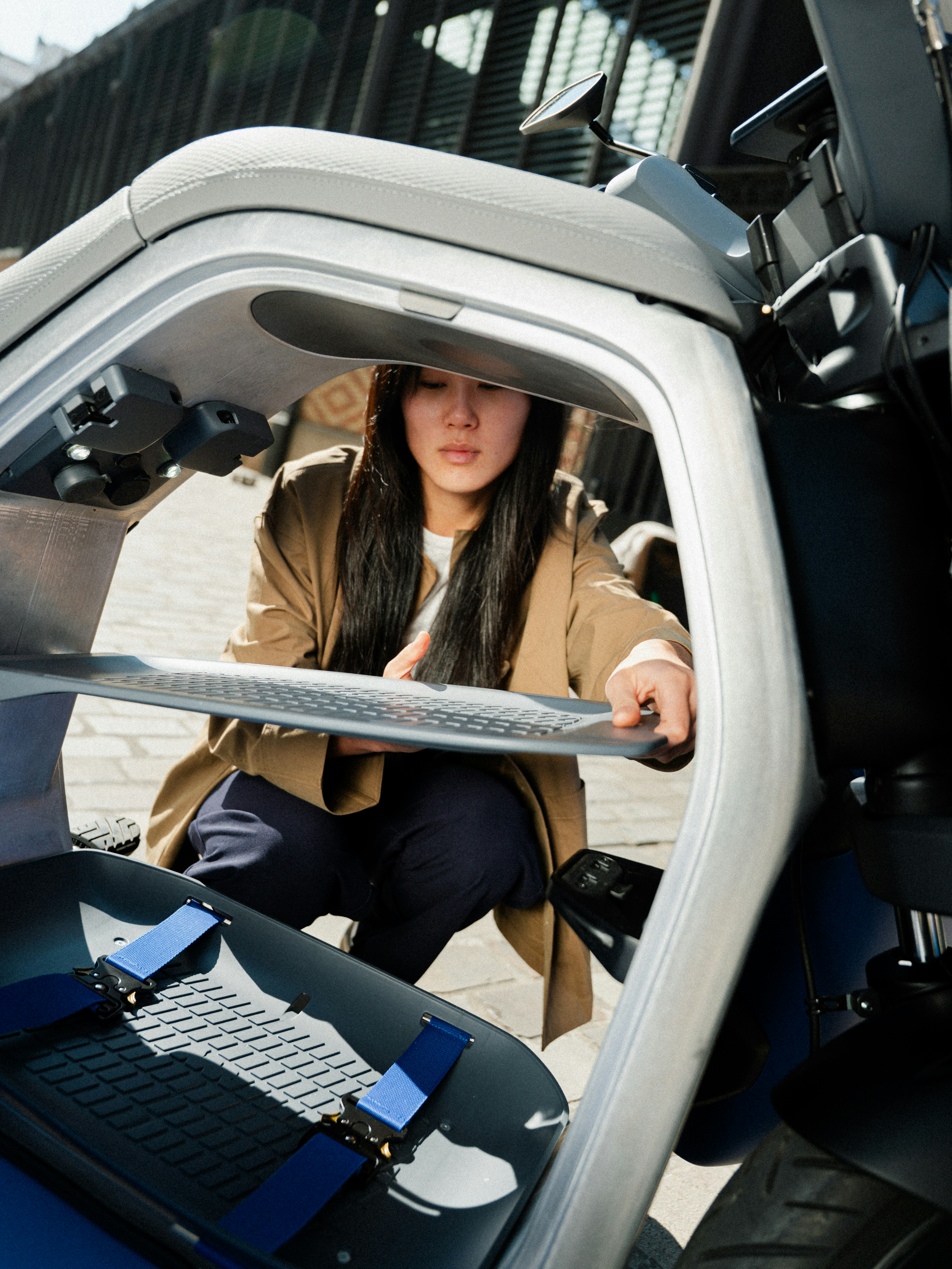 A person kneels outside an open urban vehicle, viewing its interior, with seatbelts visible. Urban setting in the background.