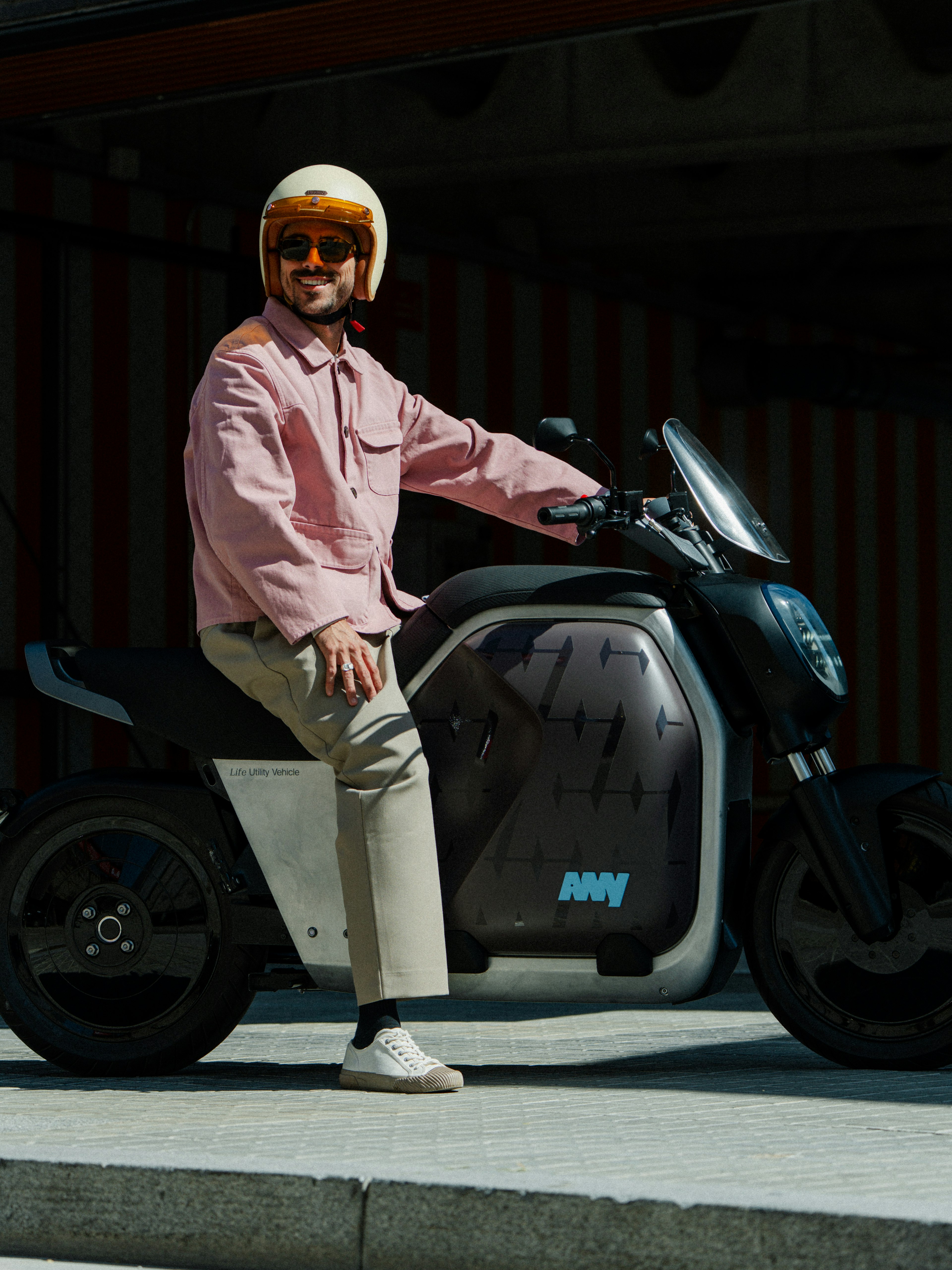 Uomo in giacca rosa e pantaloni beige, con casco, seduto su uno scooter elettrico scuro all'aria aperta.