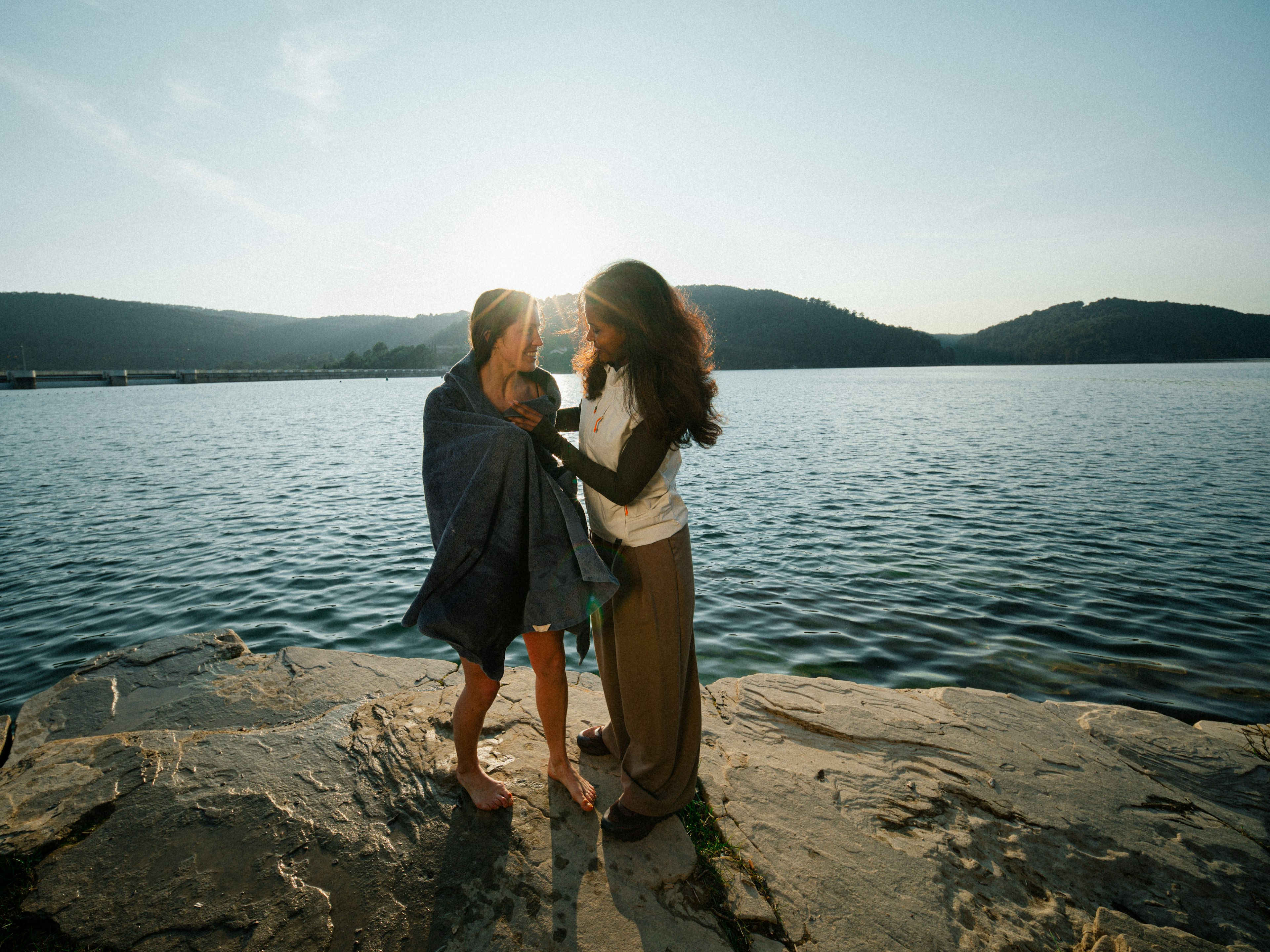 Due persone in piedi su una roccia in riva a un lago, avvolte in una coperta, con colline e un cielo luminoso sullo sfondo.
