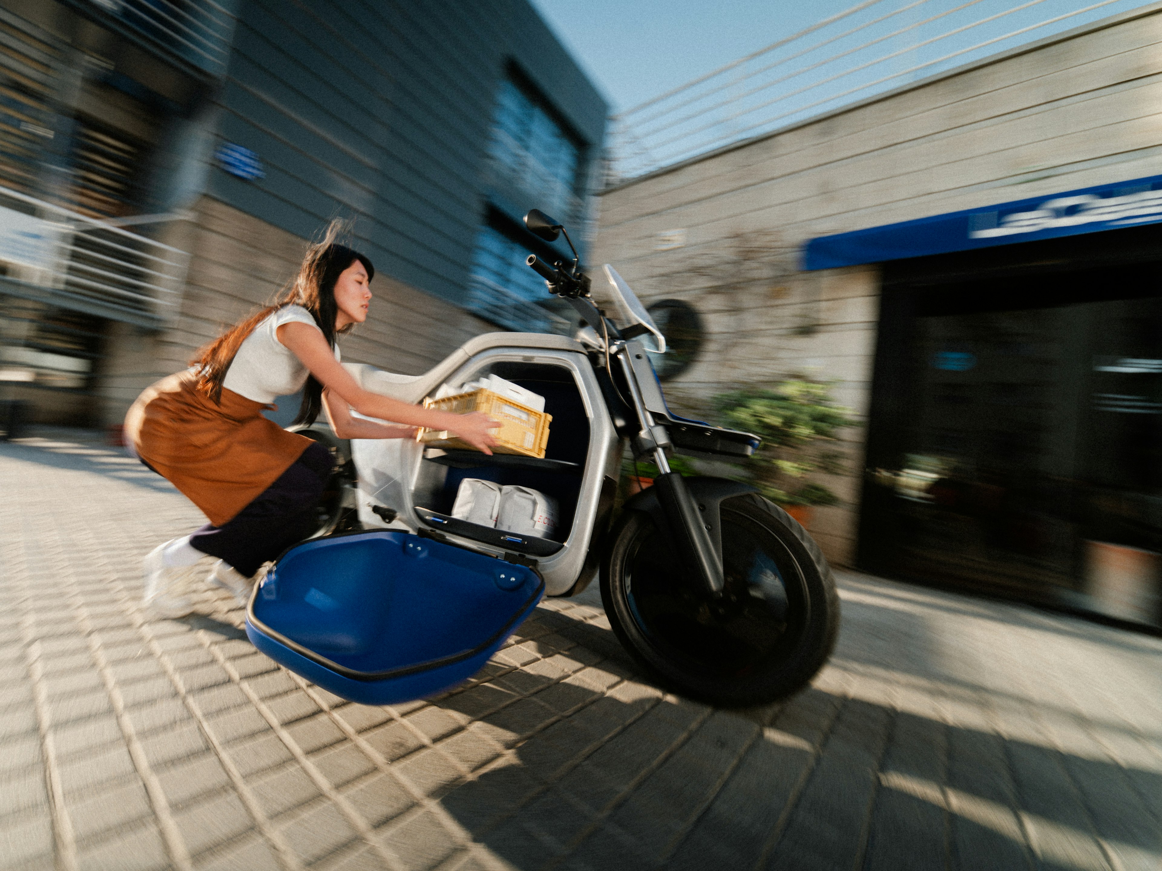 Una persona su un'elegante moto elettrica, con effetto sfocatura da movimento, con una borsa in mano, che passa davanti a un edificio moderno.
