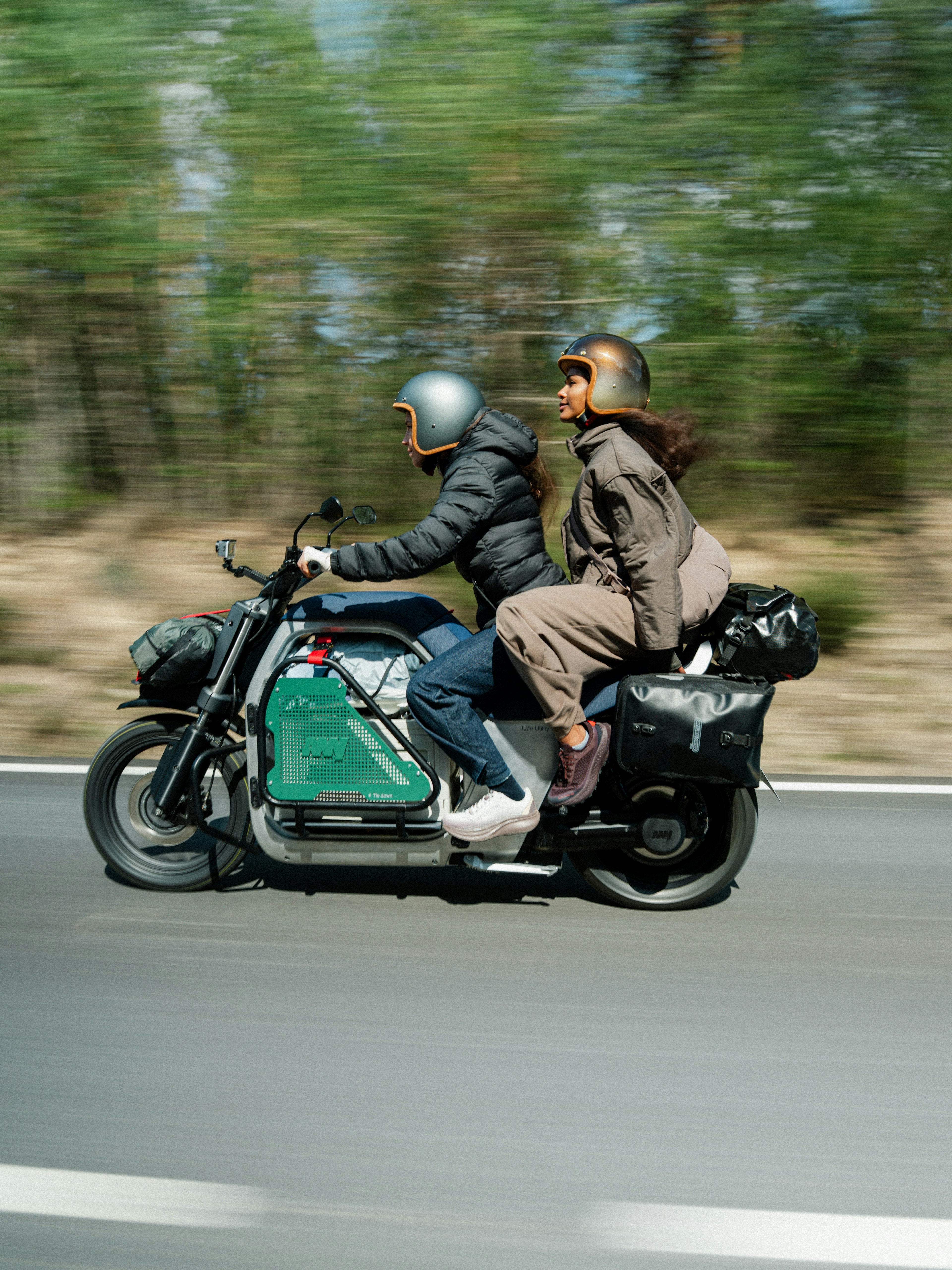Two people riding a motorcycle on a road, both wearing helmets; blurred background indicates motion.