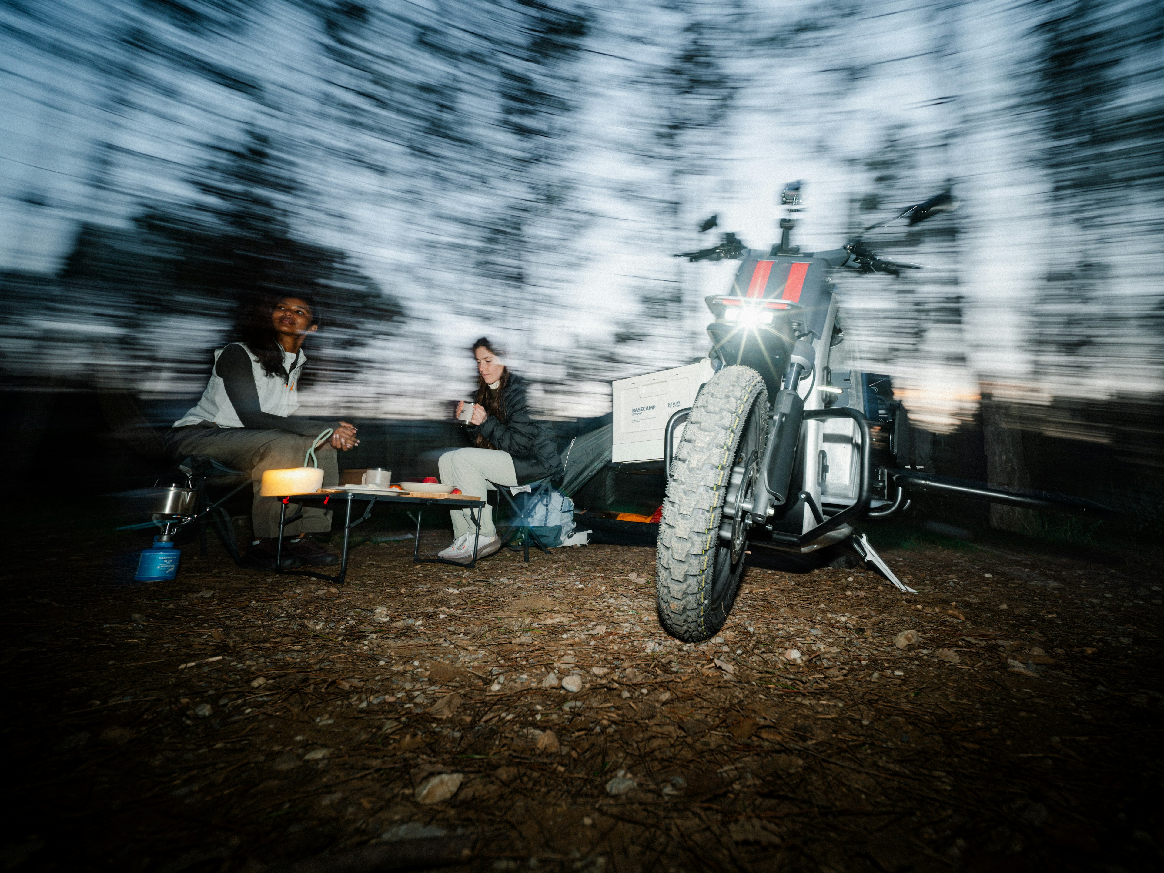Una moto parcheggiata accanto a un falò; due persone sedute su una panchina con alberi sfumati sullo sfondo, che suggeriscono movimento o vento.