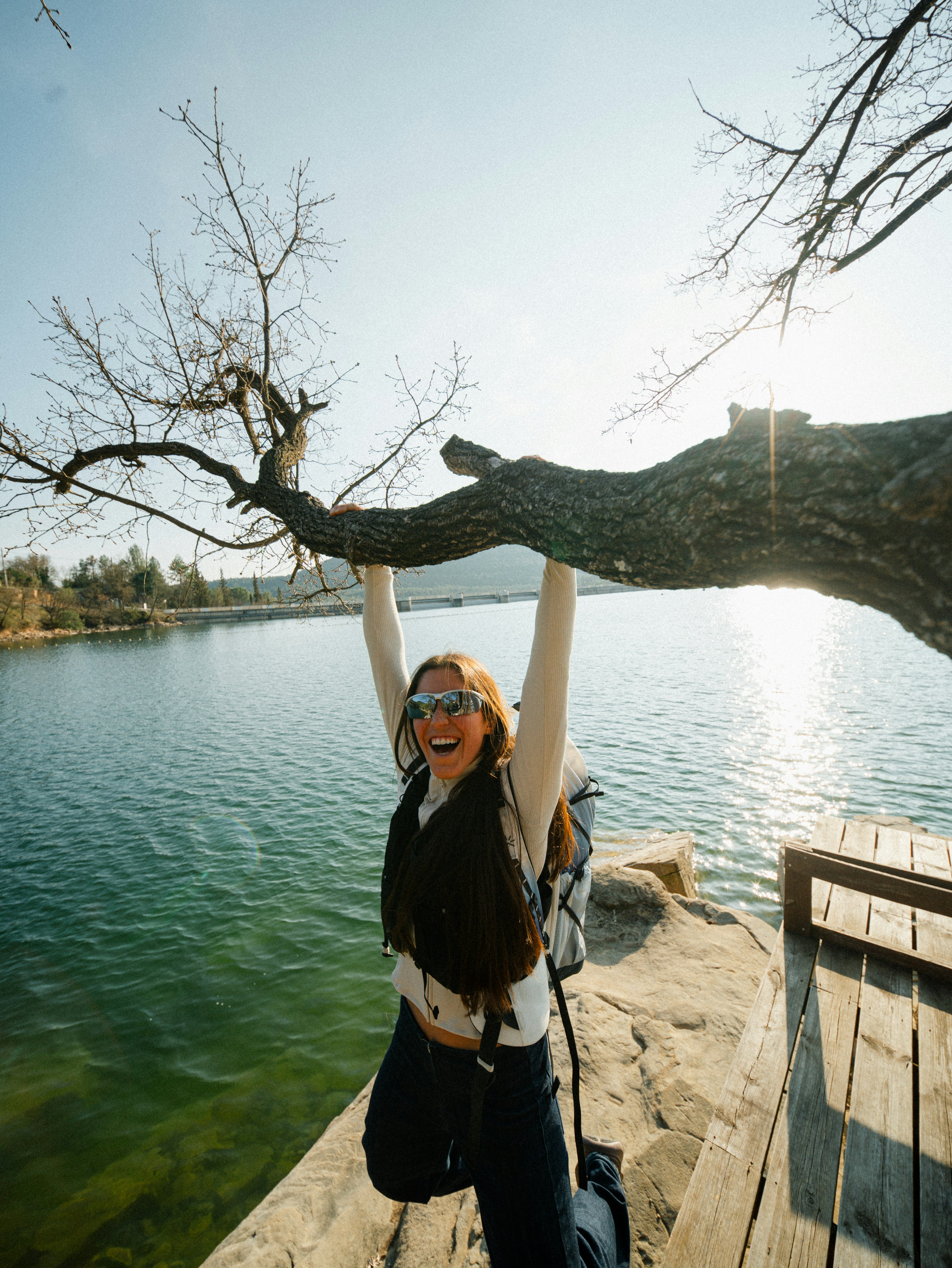 Una persona si appende al ramo di un albero in riva a un lago, sorridendo, mentre la luce del sole si riflette sull'acqua.