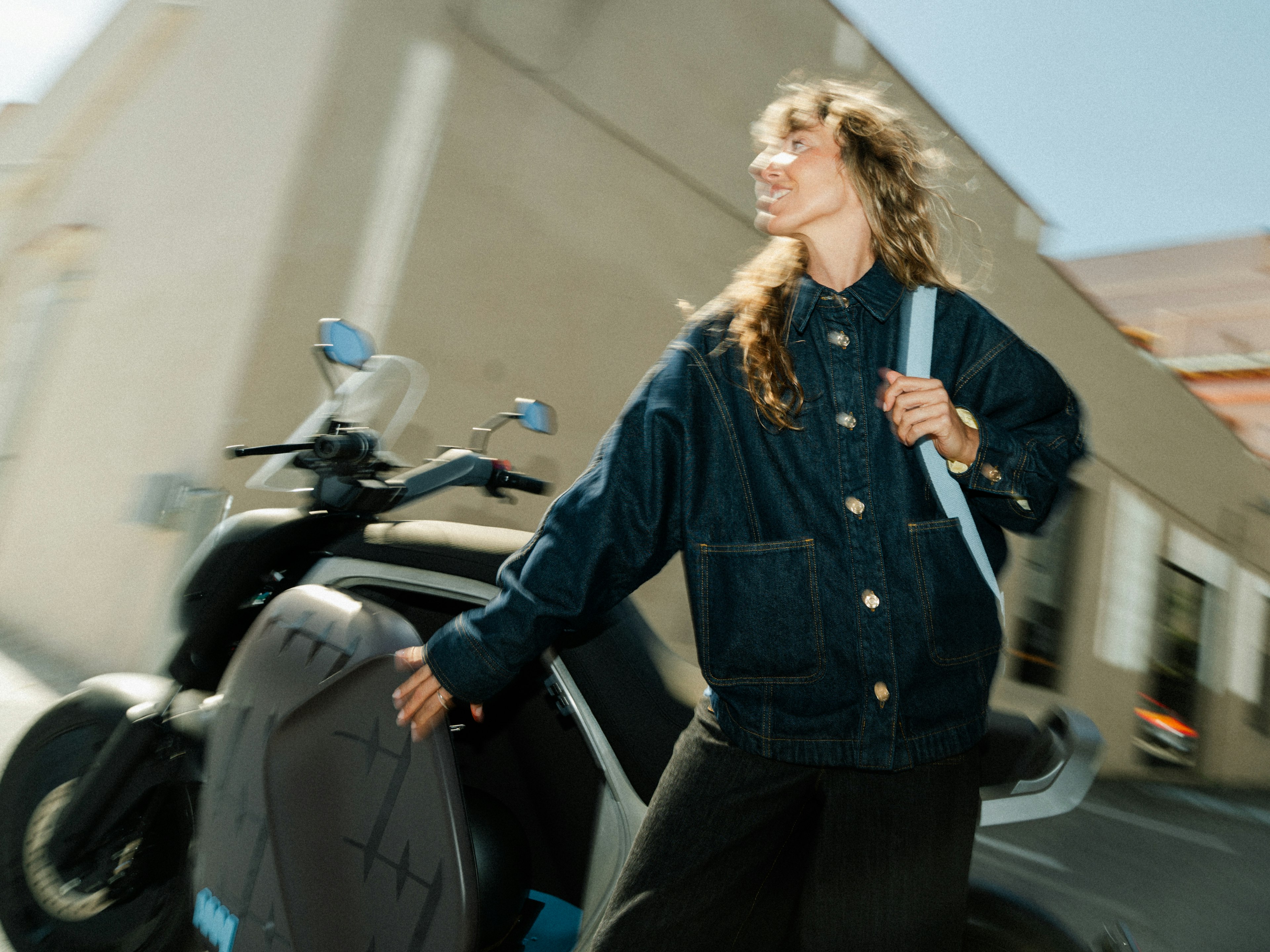 Una mujer con chaqueta vaquera de pie junto a una moto aparcada en un día soleado, sujetando el manillar y mirando al frente.