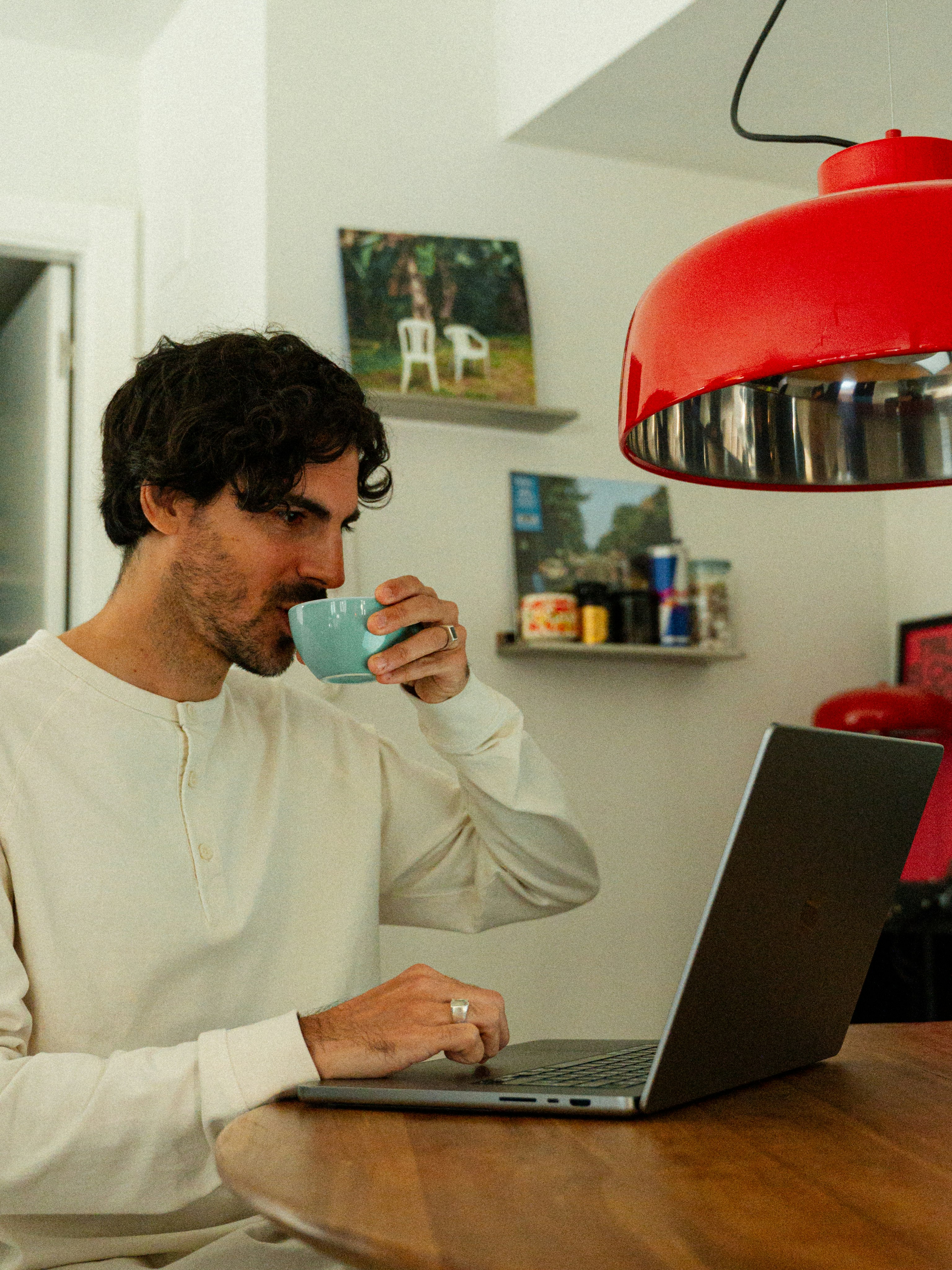 Un hombre con camisa blanca tomando café mientras trabaja con un ordenador portátil en una mesa de madera bajo una lámpara roja en una habitación acogedora.