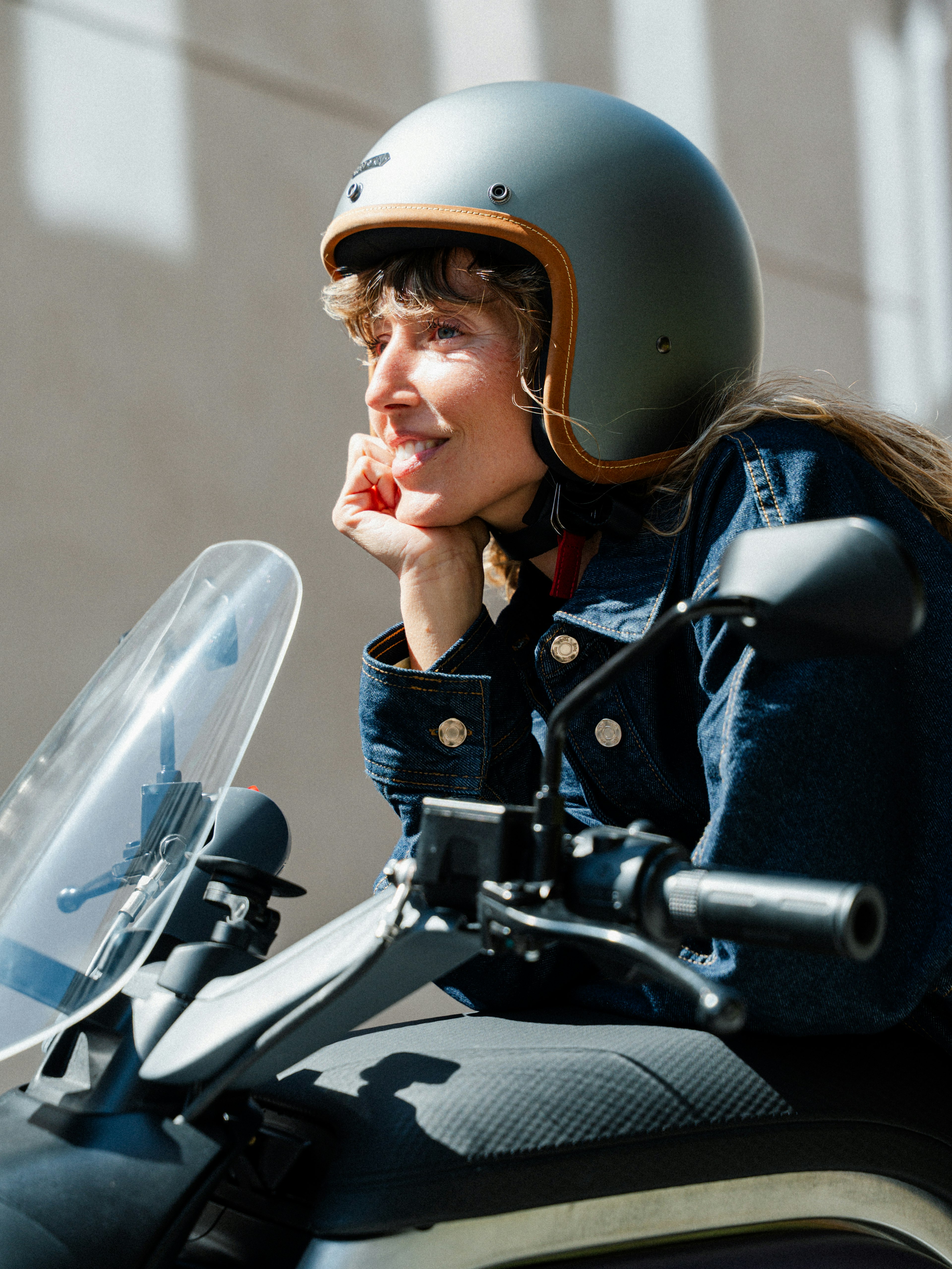 Una mujer sonriente con casco apoya la barbilla en la mano, sentada en una motocicleta con parabrisas transparente.
