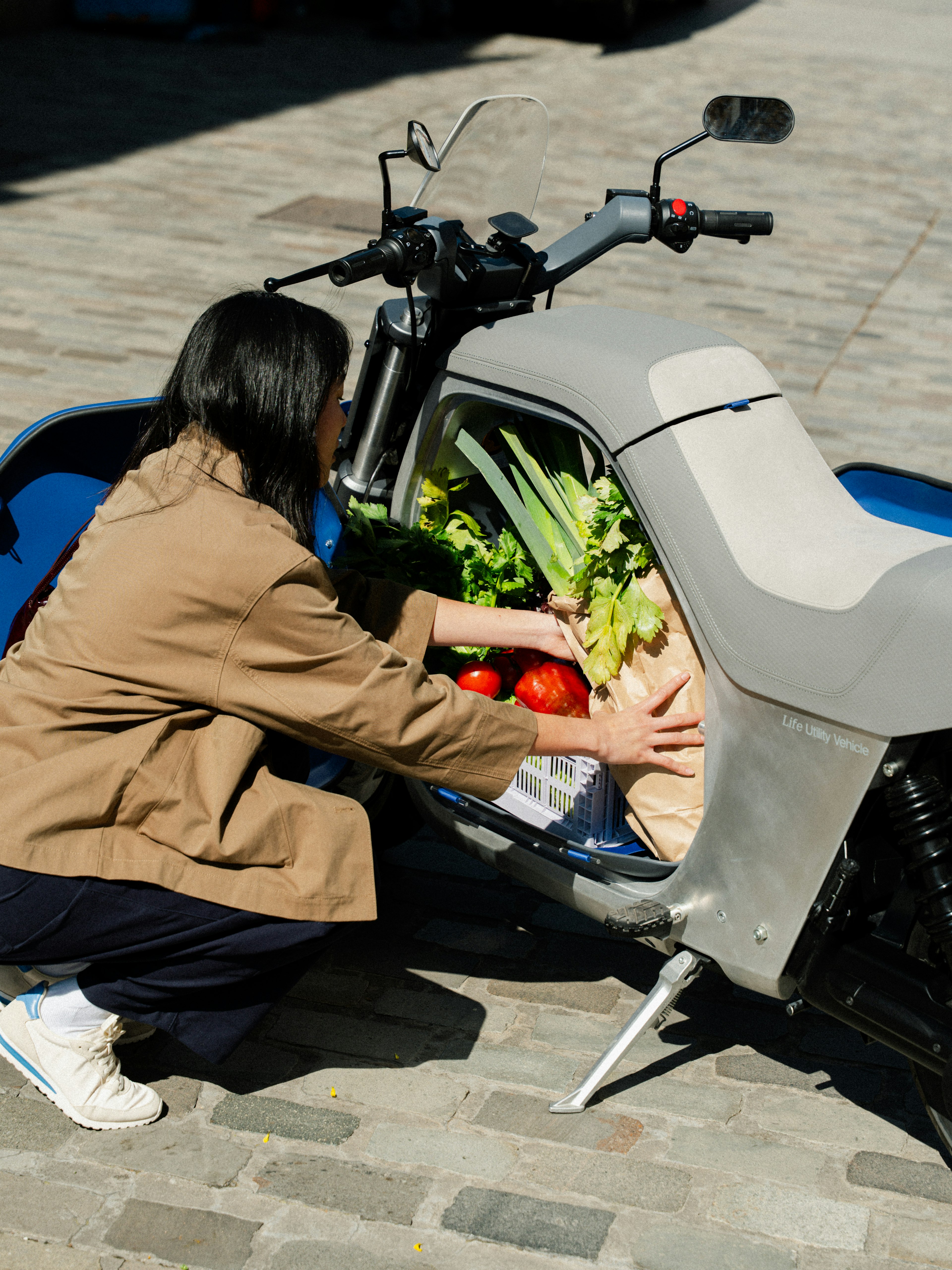 Una persona si inginocchia per riporre alcuni generi alimentari, tra cui sedano e pomodori, in un vano di uno scooter blu parcheggiato su una strada acciottolata.