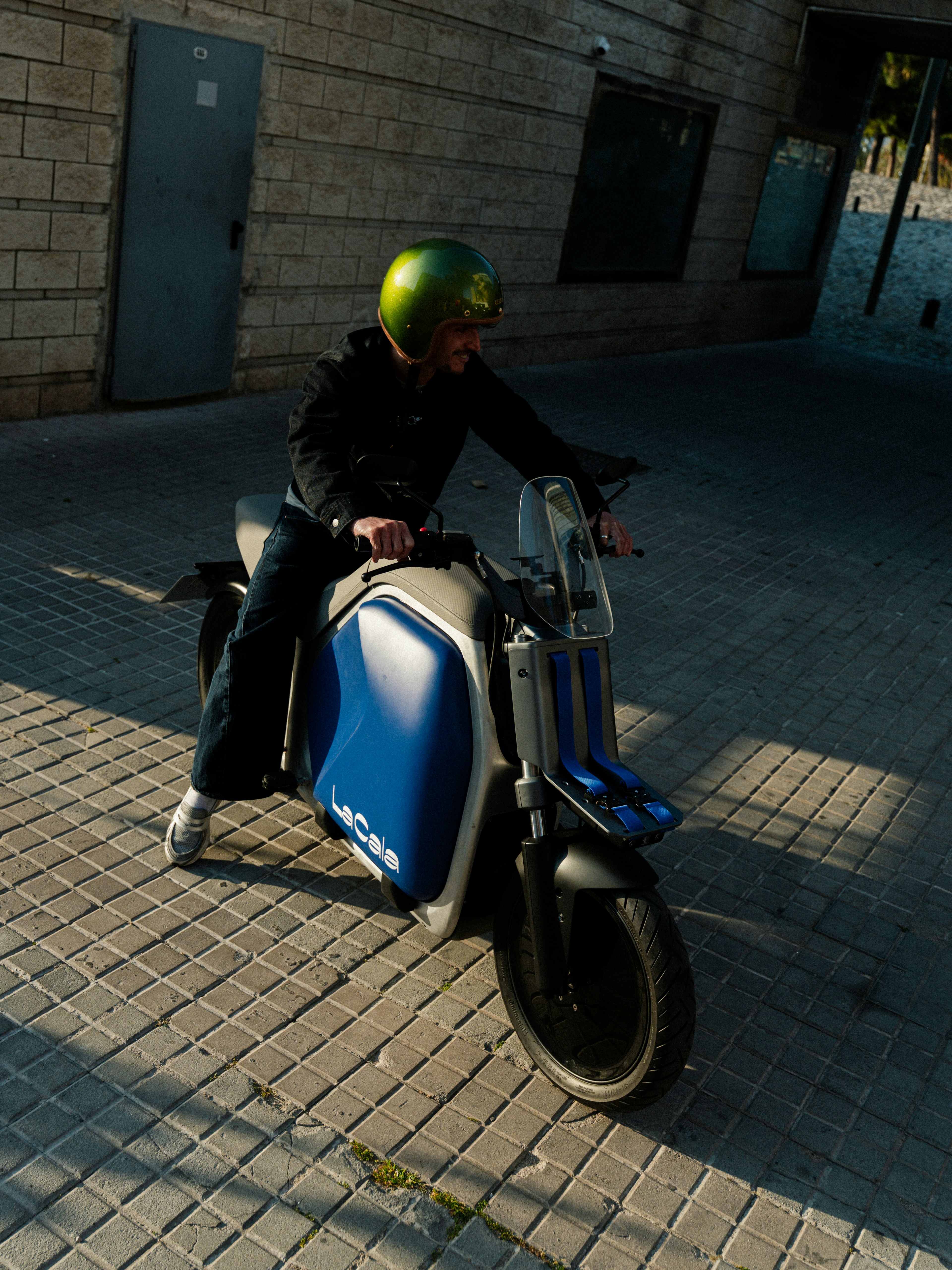 Una persona con un casco verde in sella a uno scooter blu su un sentiero di mattoni illuminato dal sole.