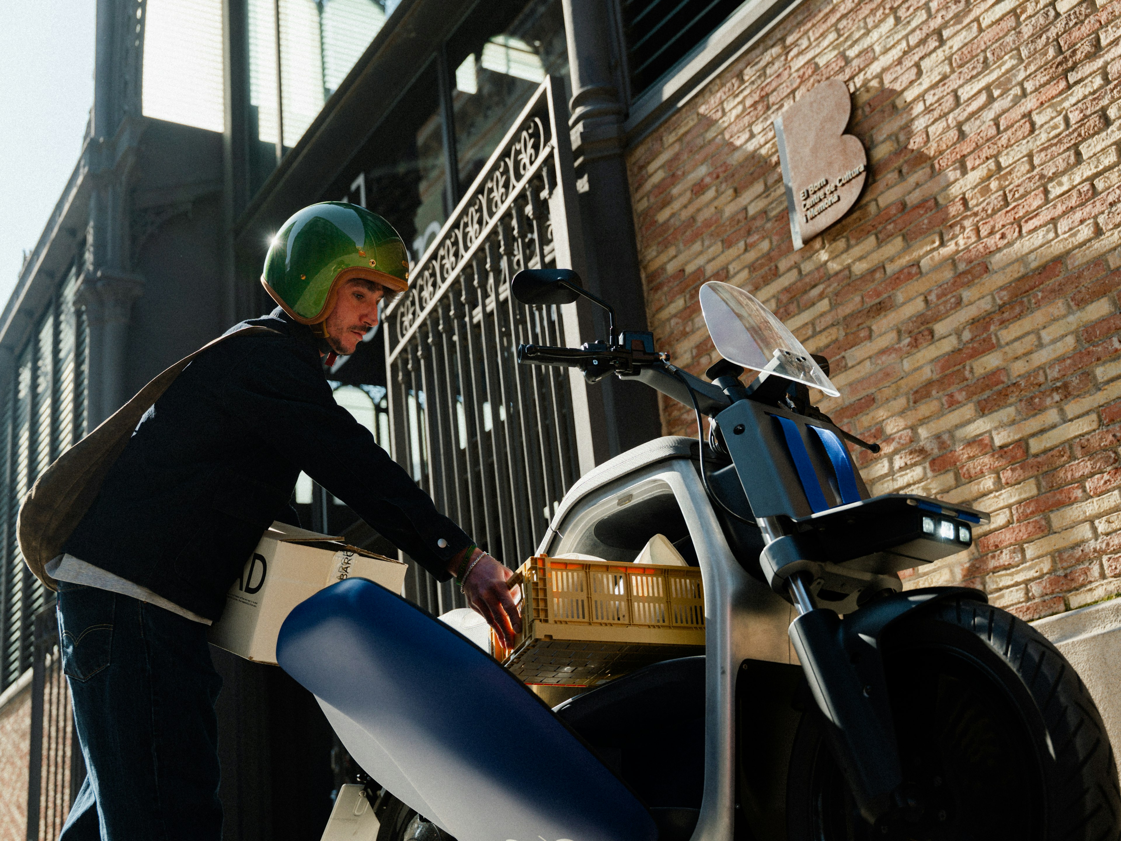 Un uomo con un casco verde appoggia un cestino su una moto parcheggiata davanti a un muro di mattoni su cui è affisso un cartello astratto.
