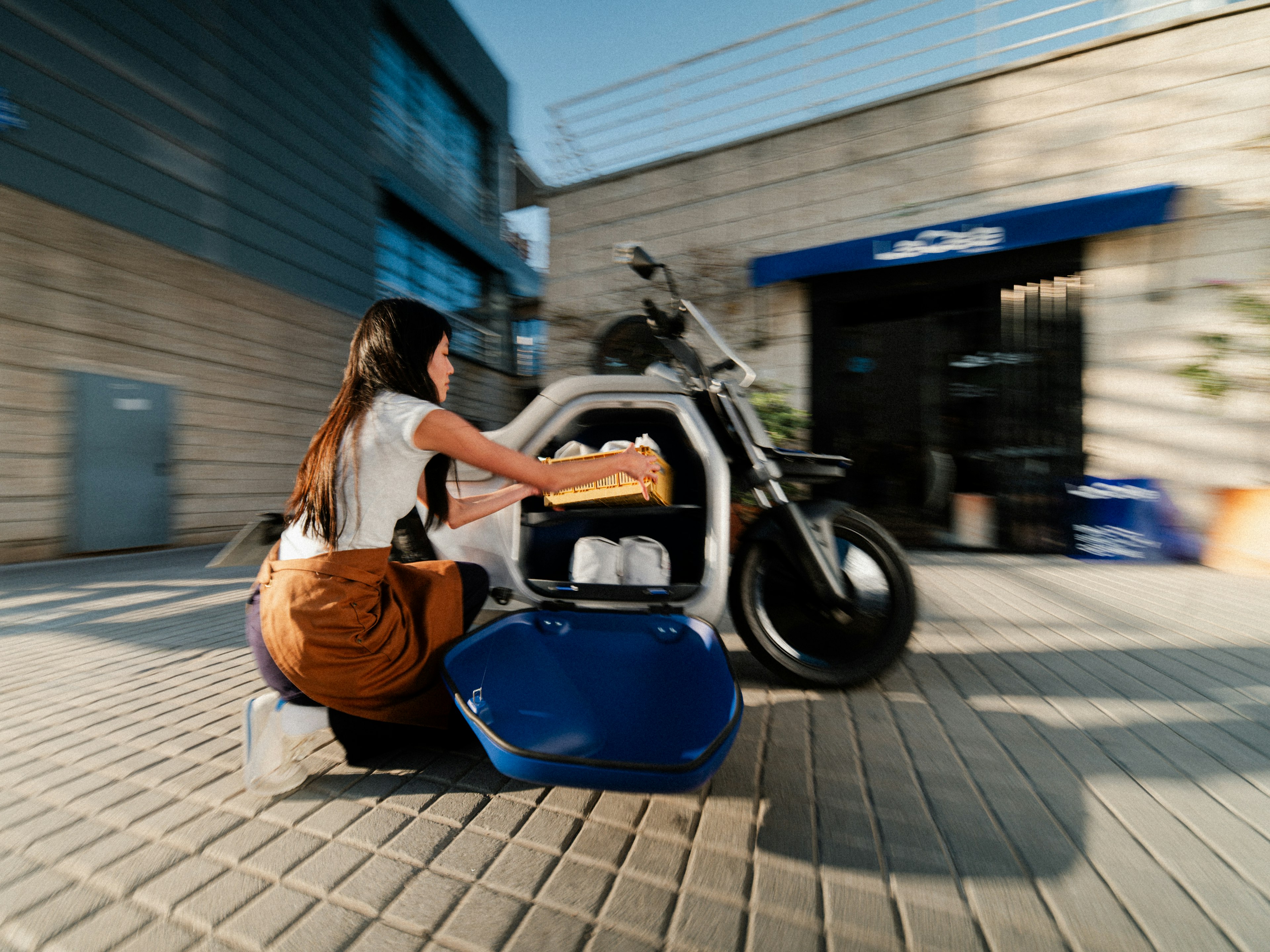 A woman with a backpack examines a motorcycle parked on a sunlit street beside a brick building.