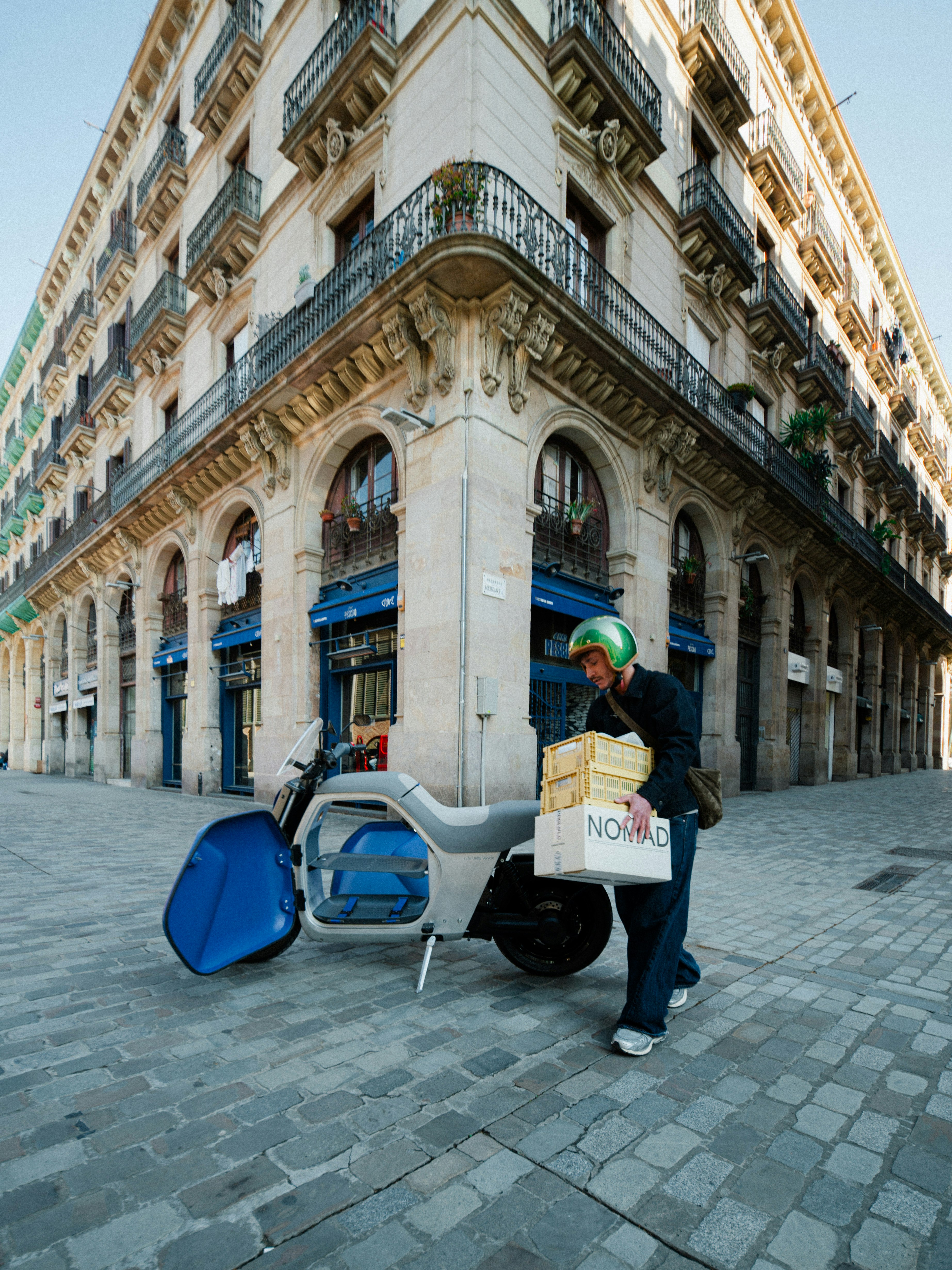 Un uomo che trasporta dei pacchi accanto a un rimorchio per biciclette su una strada acciottolata, davanti a un edificio storico con finestre ad arco.