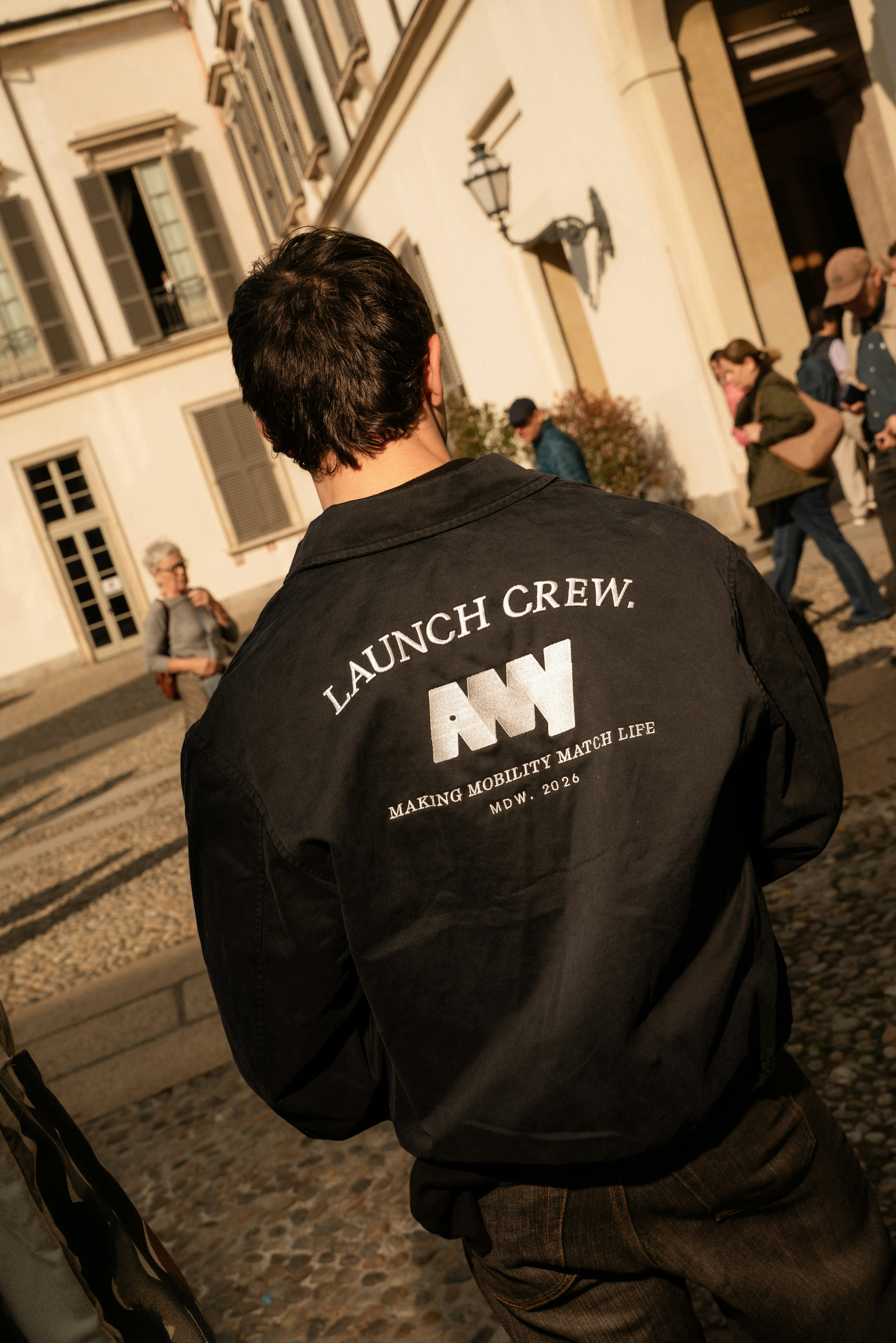 Man in "LAUNCH CREW" jacket walks outdoors, sunny day, with historic buildings and people in the background.