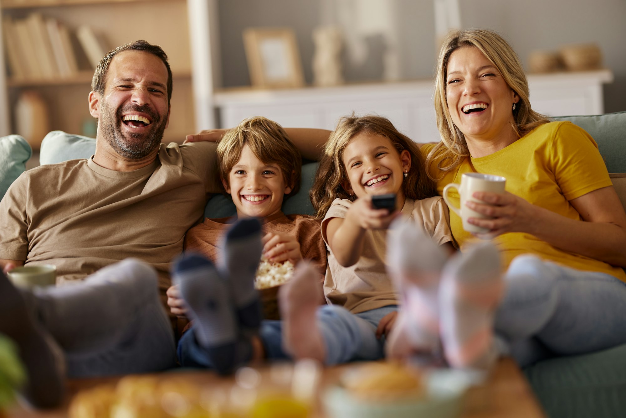 4 Köpfige junge Familie sitzt vergnügt auf Sofa und schaut TV.