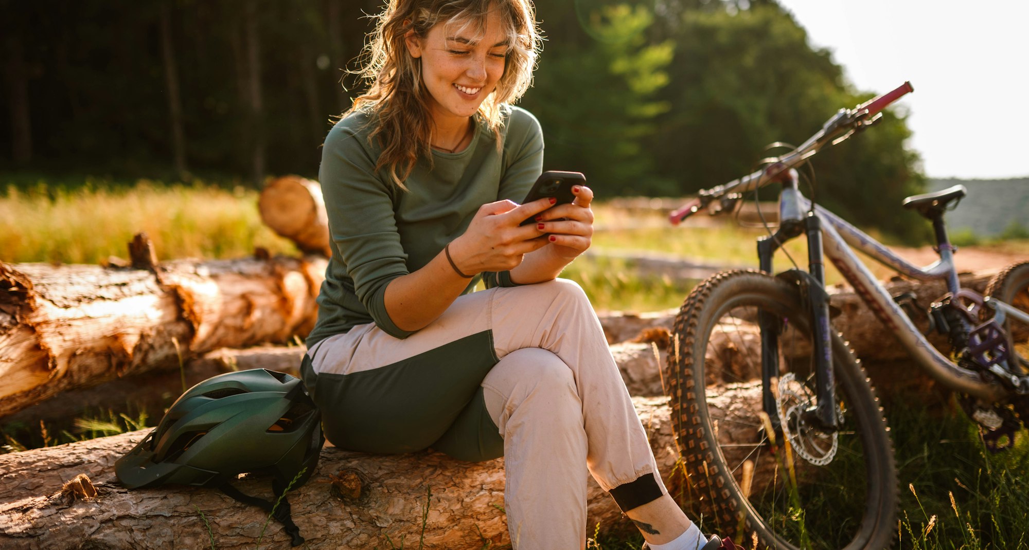 Junge Bikerin sitzt neben ihrem Fahrrad in der Natur und blickt auf ihr Smartphone.