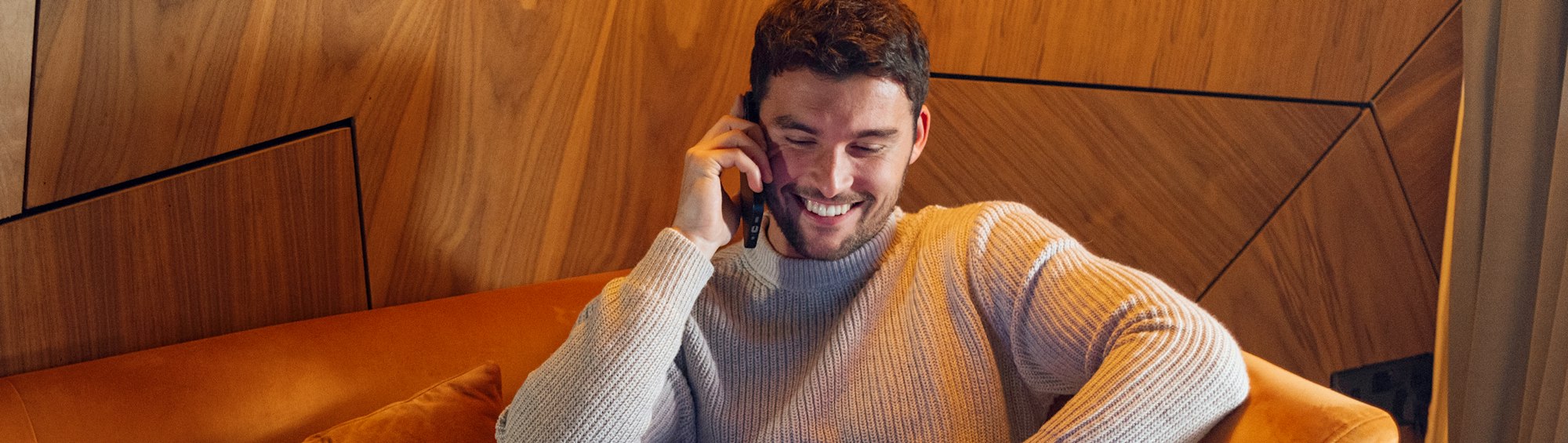 Lächelnder Mann im Strickpullover telefoniert entspannt auf einem orangefarbenen Sofa vor einer Holzwand.