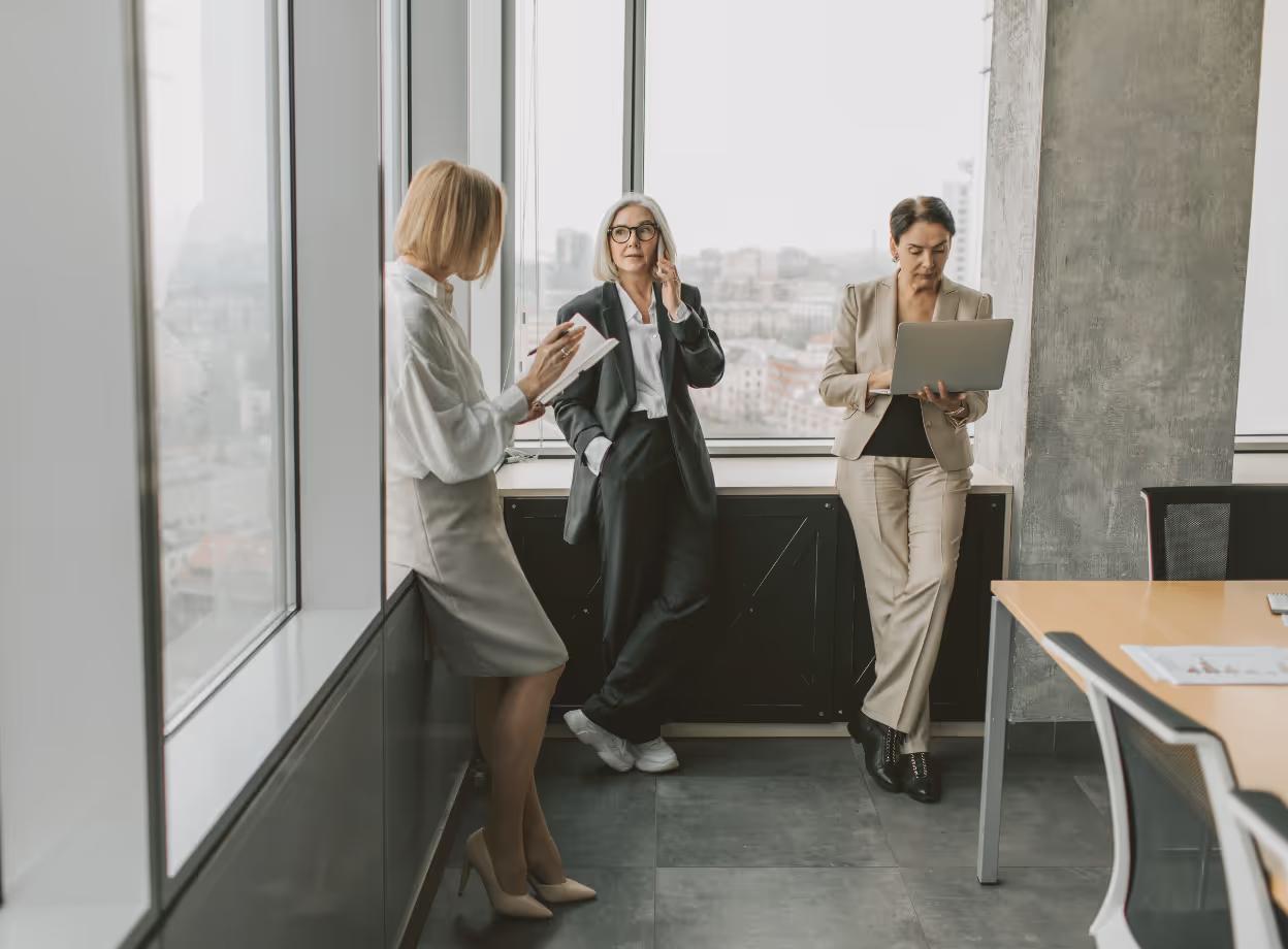 Drei Geschäftsfrauen in modernem Büro besprechen flexible Arbeitszeitmodelle und Brückenteilzeit für bessere Work-Life-Balance.