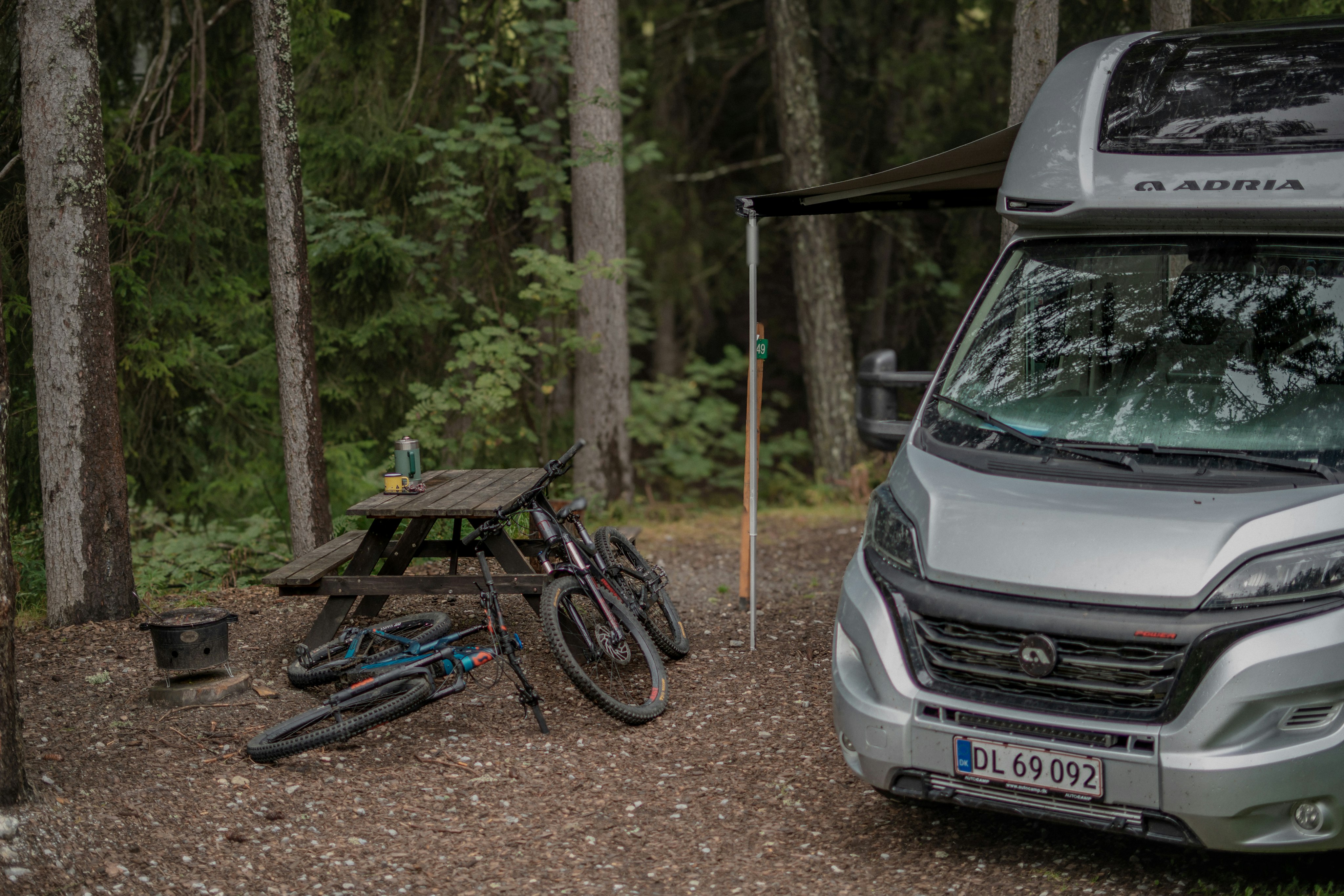 Grauer Campervan mit Vordach und zwei Bikes im Tannenwald auf dem Campingplatz.