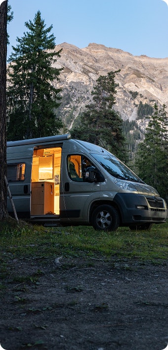 Beleuchteter Campervan bei Dämmerung vor Bergkulisse und Tannenwald.