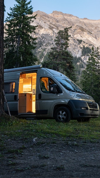Campervan am Abend Beleuchteter Campervan bei Dämmerung vor Bergkulisse und Tannenwald.