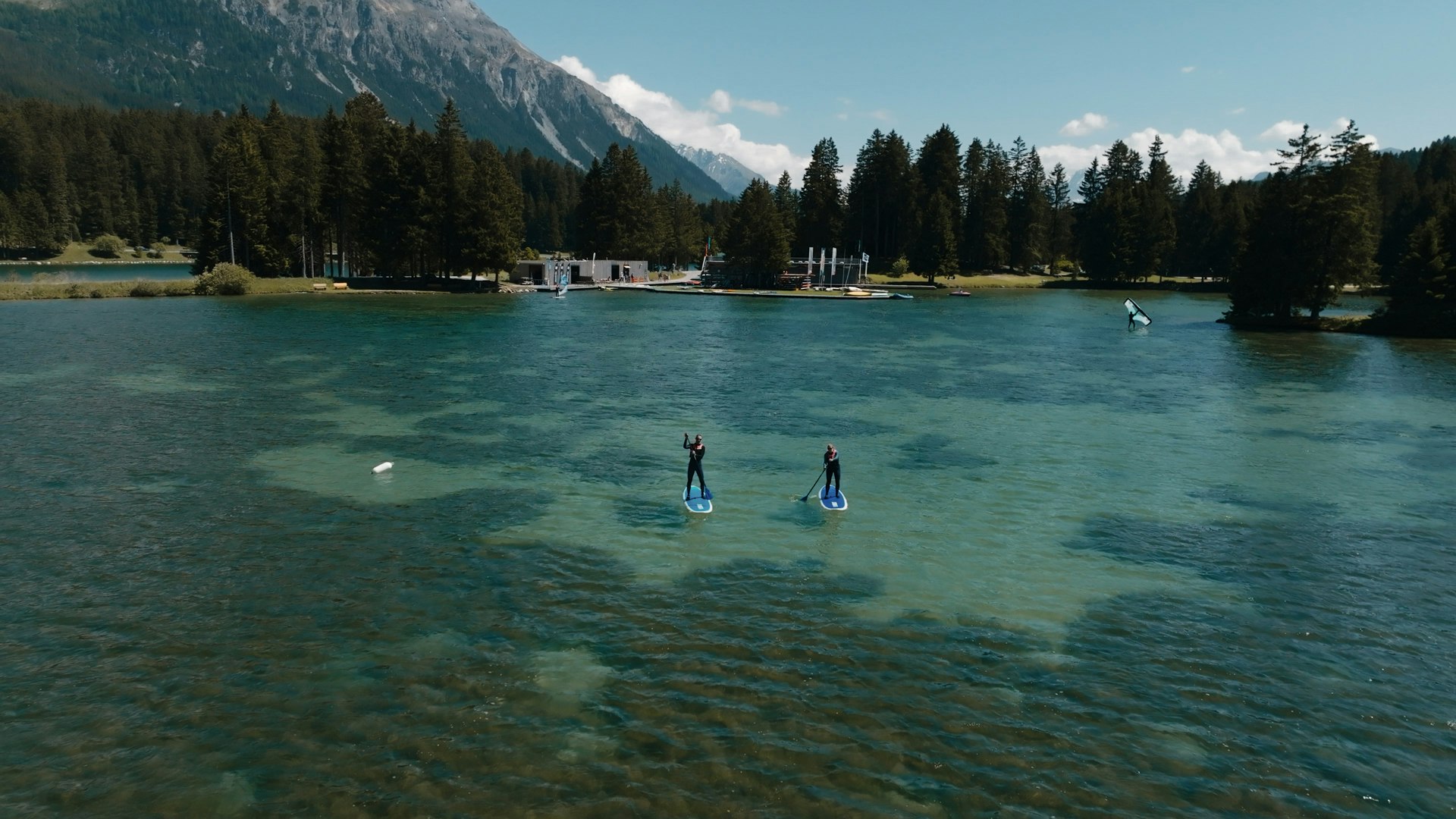 Klarer Bergsee umgeben von dichtem Nadelwald und SUPs