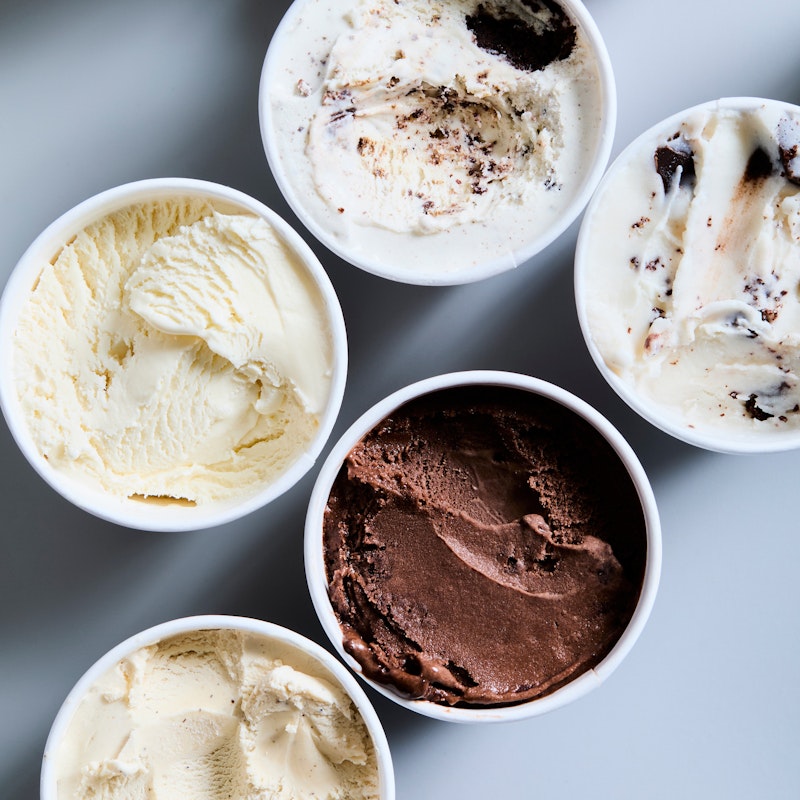 Seven tubs of Village Ice Cream arranged in two vertical columns, showing various flavours including vanilla, chocolate, and cookies and cream, with textured scoops visible on the surface in each tub.