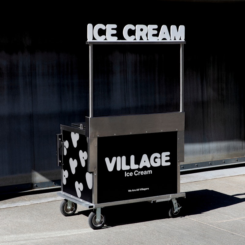 A black and white ice cream cart with VILLAGE Ice Cream on the side and ICE CREAM on top stands outside near a brick wall, in sunlight.