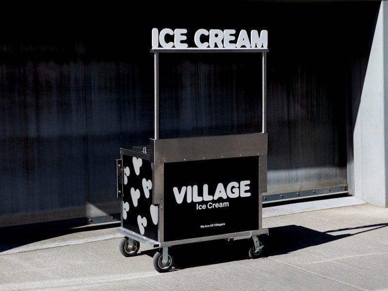 A black and white ice cream cart with VILLAGE Ice Cream on the side and ICE CREAM on top stands outside near a brick wall, in sunlight.