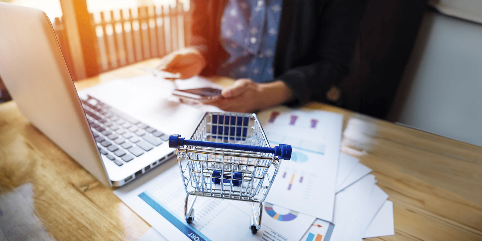 Graphic featuring a shopping cart, laptop, and papers on a desk, symbolizing ecommerce product synchronization and automation between NetSuite and WooCommerce.