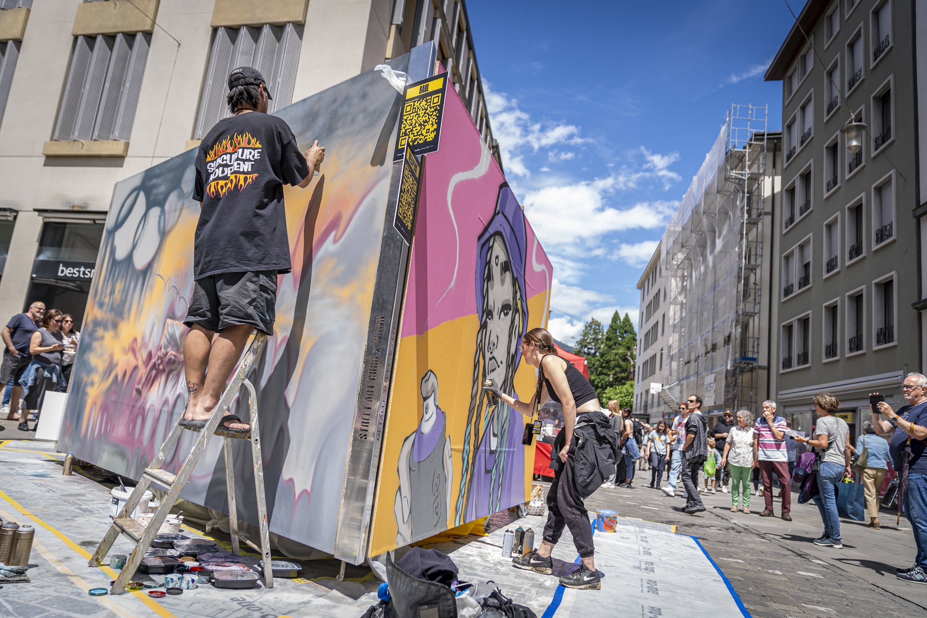A man stands on the letter with spray paint in his hand. He is working on his mural. A woman on the other side is also working on her mural. Multiple spectators are watching and taking pictures.
