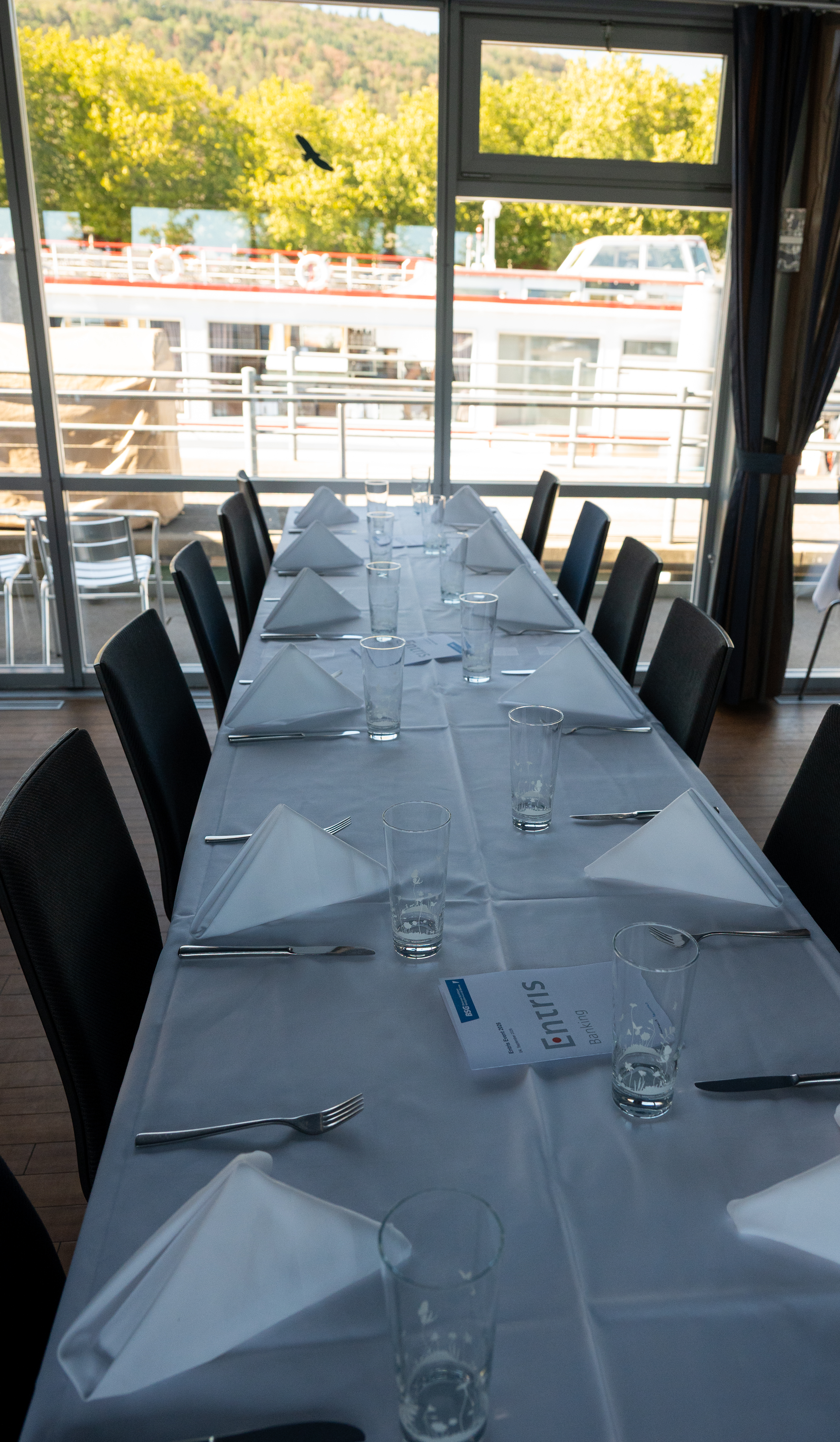 Une longue table à manger recouverte de nappes blanches, de serviettes, de verres et de couverts, avec vue sur un bateau fluvial à travers de grandes fenêtres.