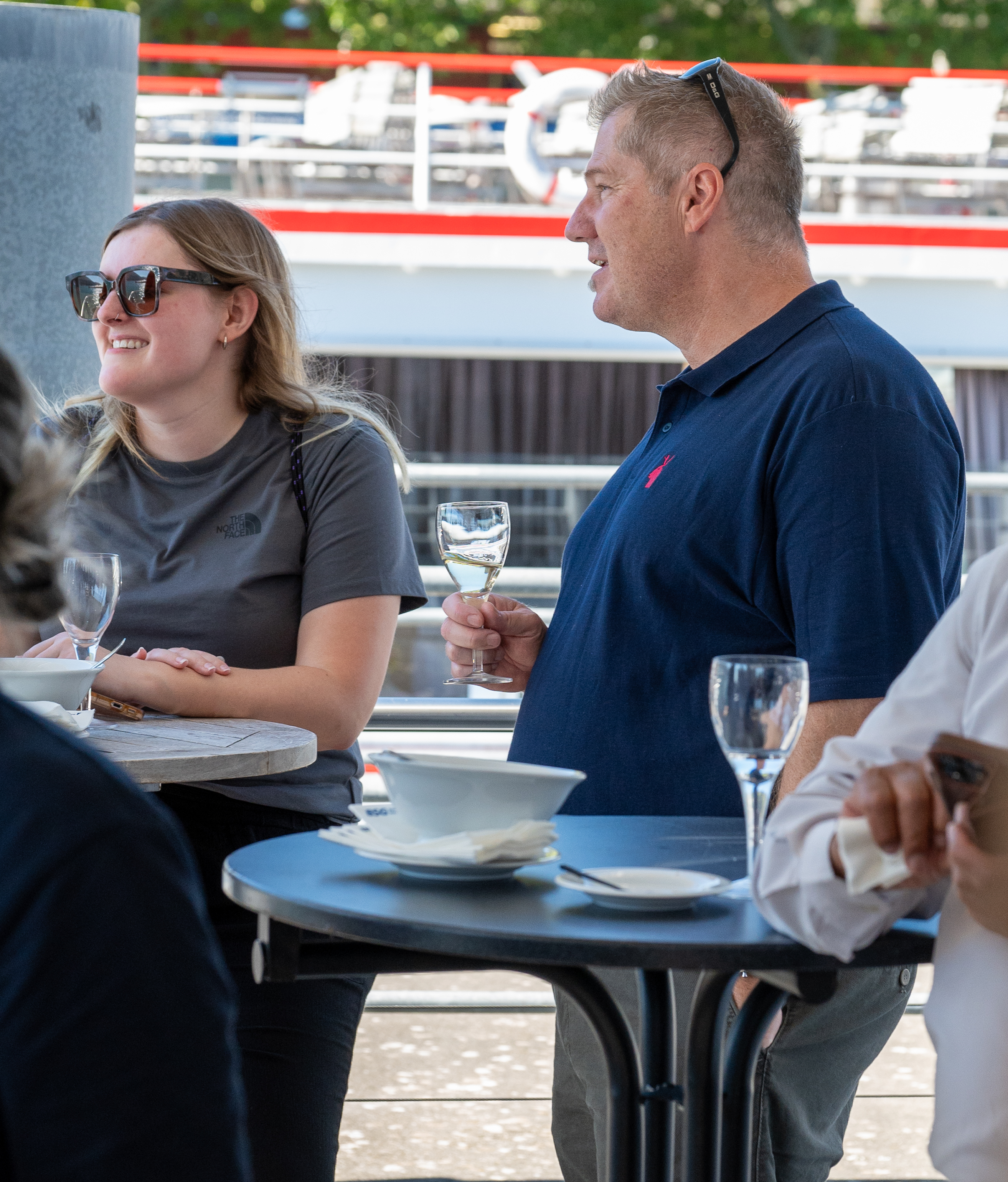 Une femme et un homme sont debout près de tables en terrasse, un verre de vin blanc à la main chacun.