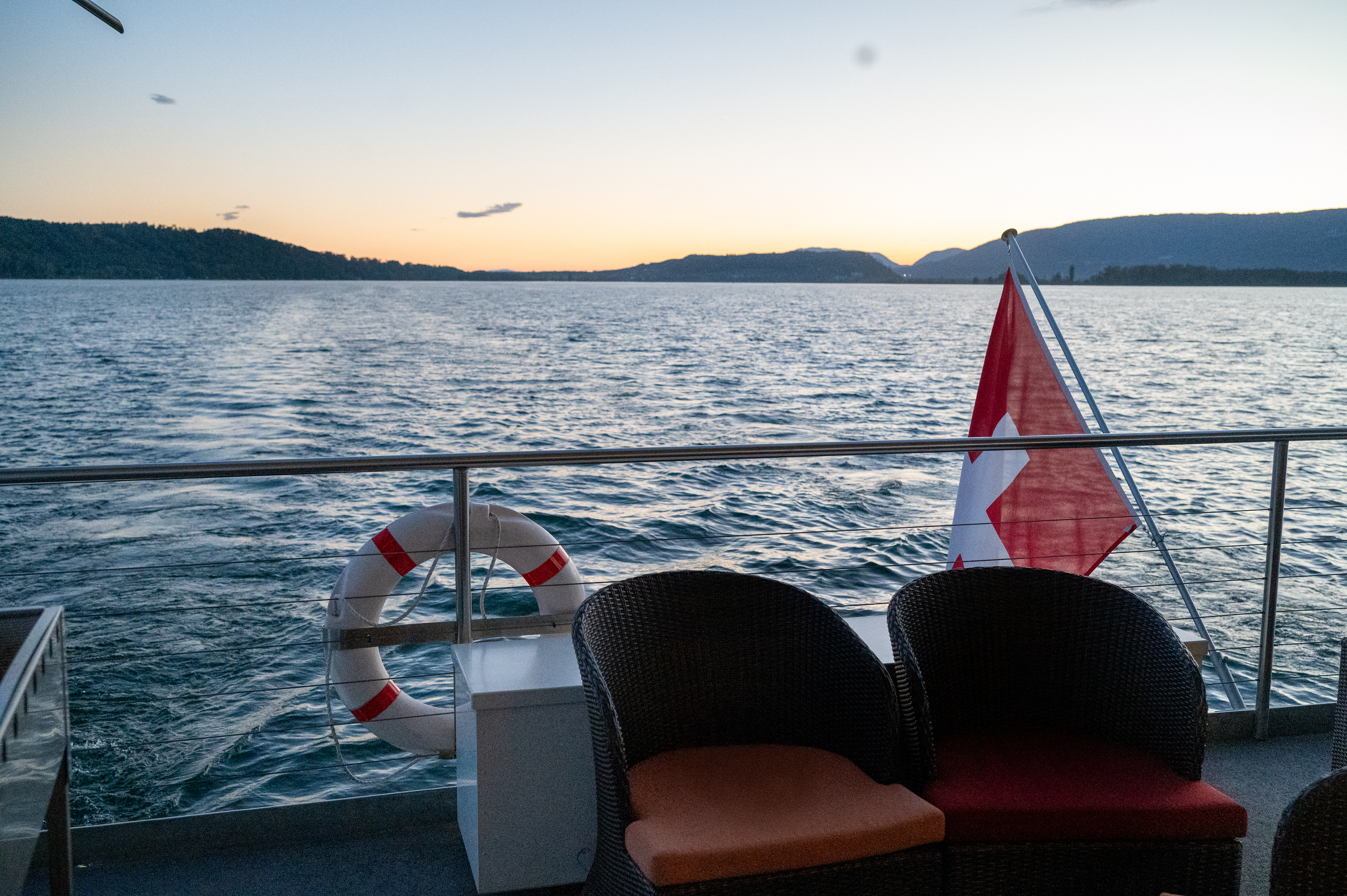Vue sur un lac depuis le pont d'un bateau, sur lequel on aperçoit une bouée de sauvetage, un drapeau suisse et deux chaises de jardin.