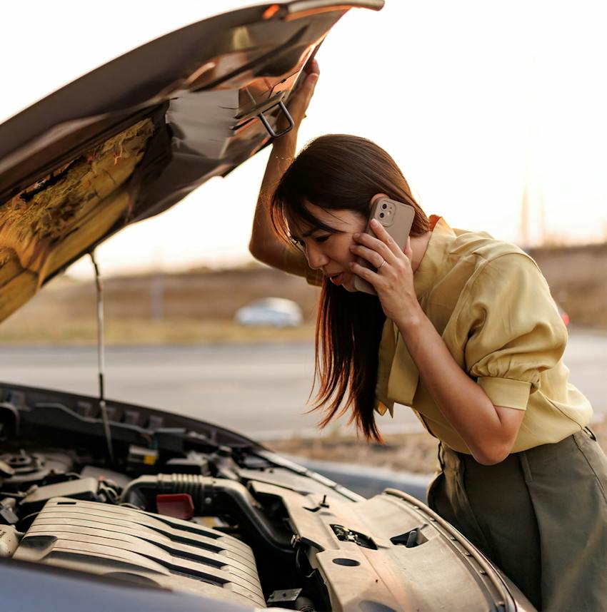 woman on the phone looking under the hood of her car