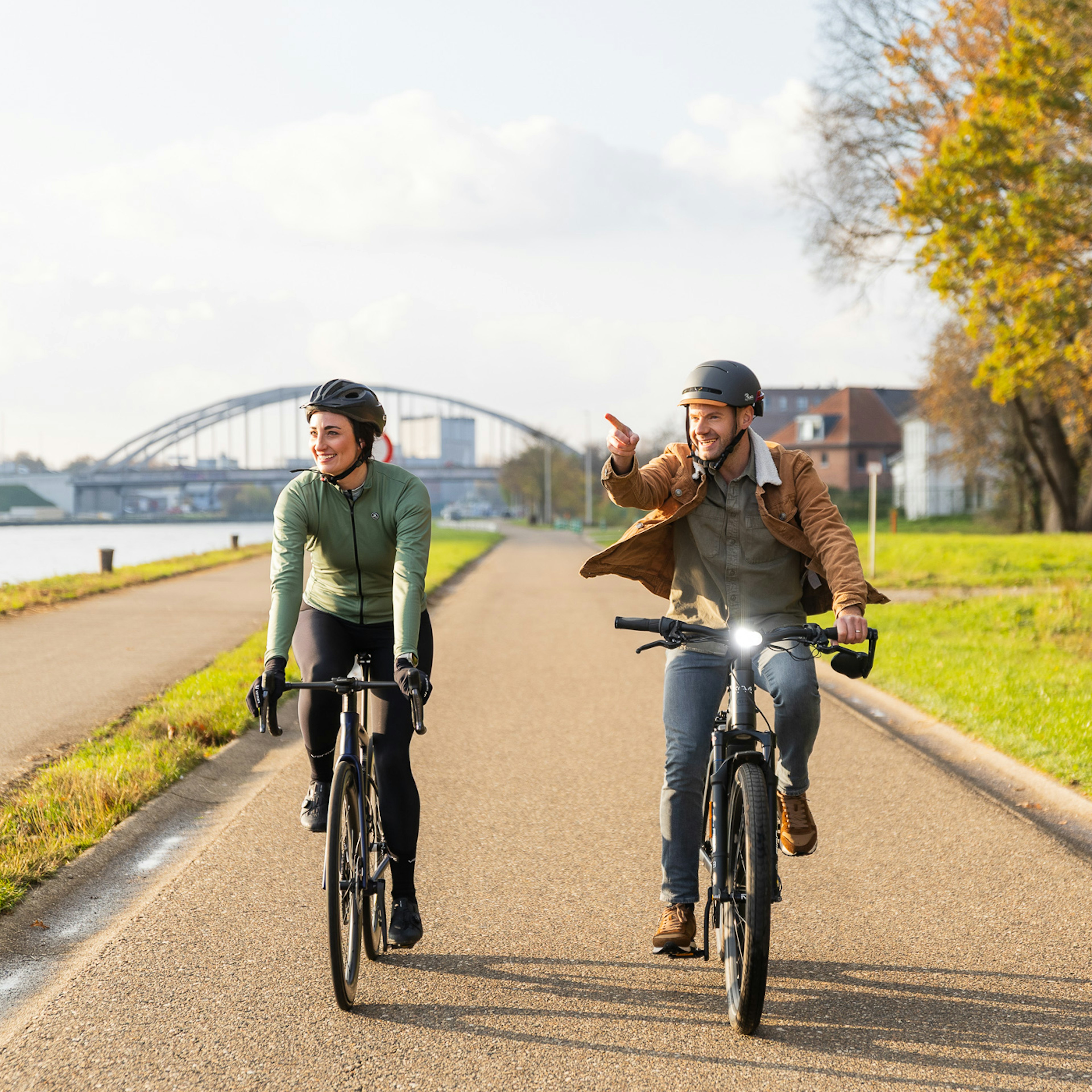 Une femme sur un vélo de route et un homme sur un vélo électrique roulent côte à côte le long d'une voie cyclable en bordure de canal baignée de soleil ; l'homme pointe du doigt devant lui, avec un pont en arc en arrière-plan.