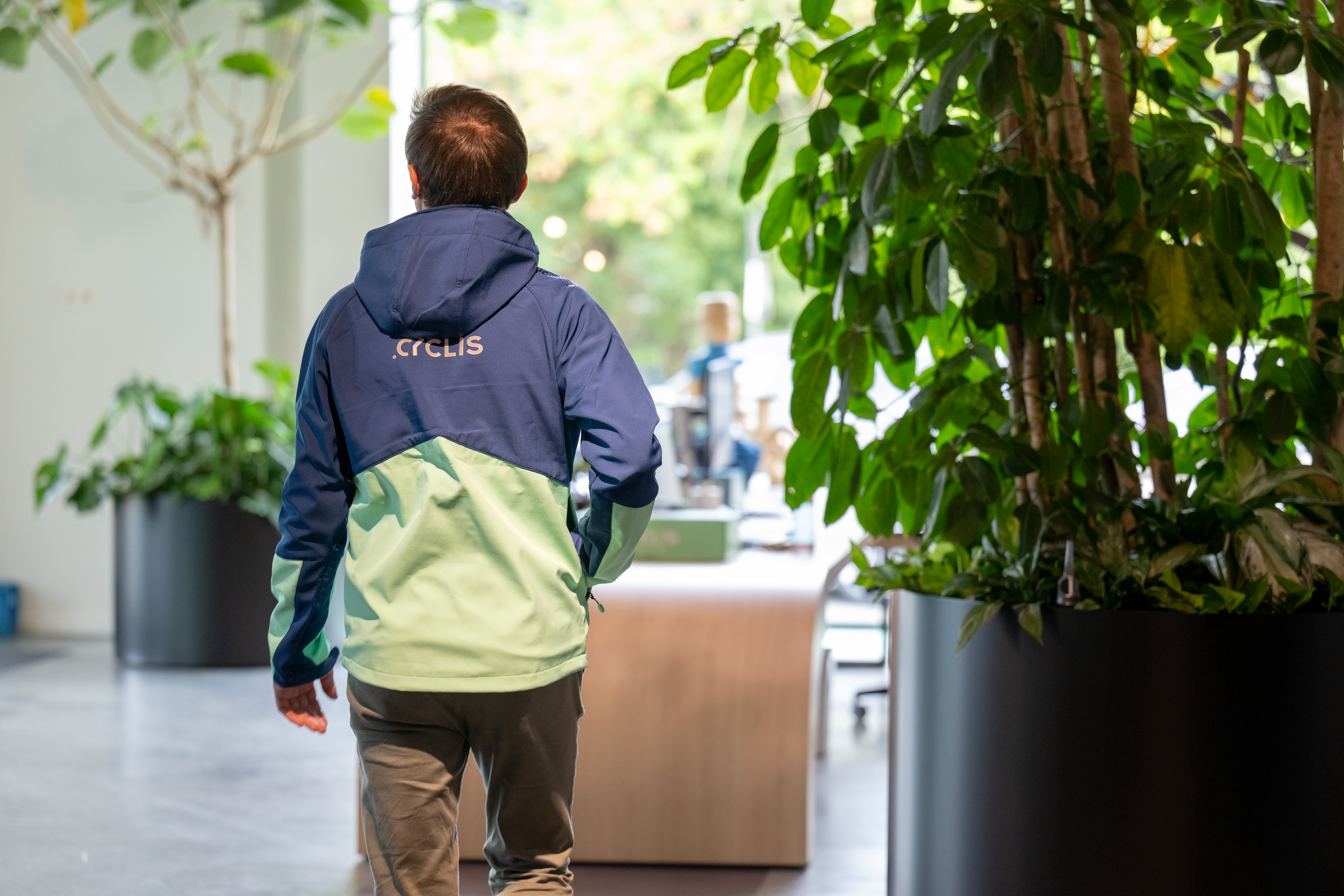 A Cyclis employee in a branded jacket walks away from the camera through the planted, daylit office toward the reception area.