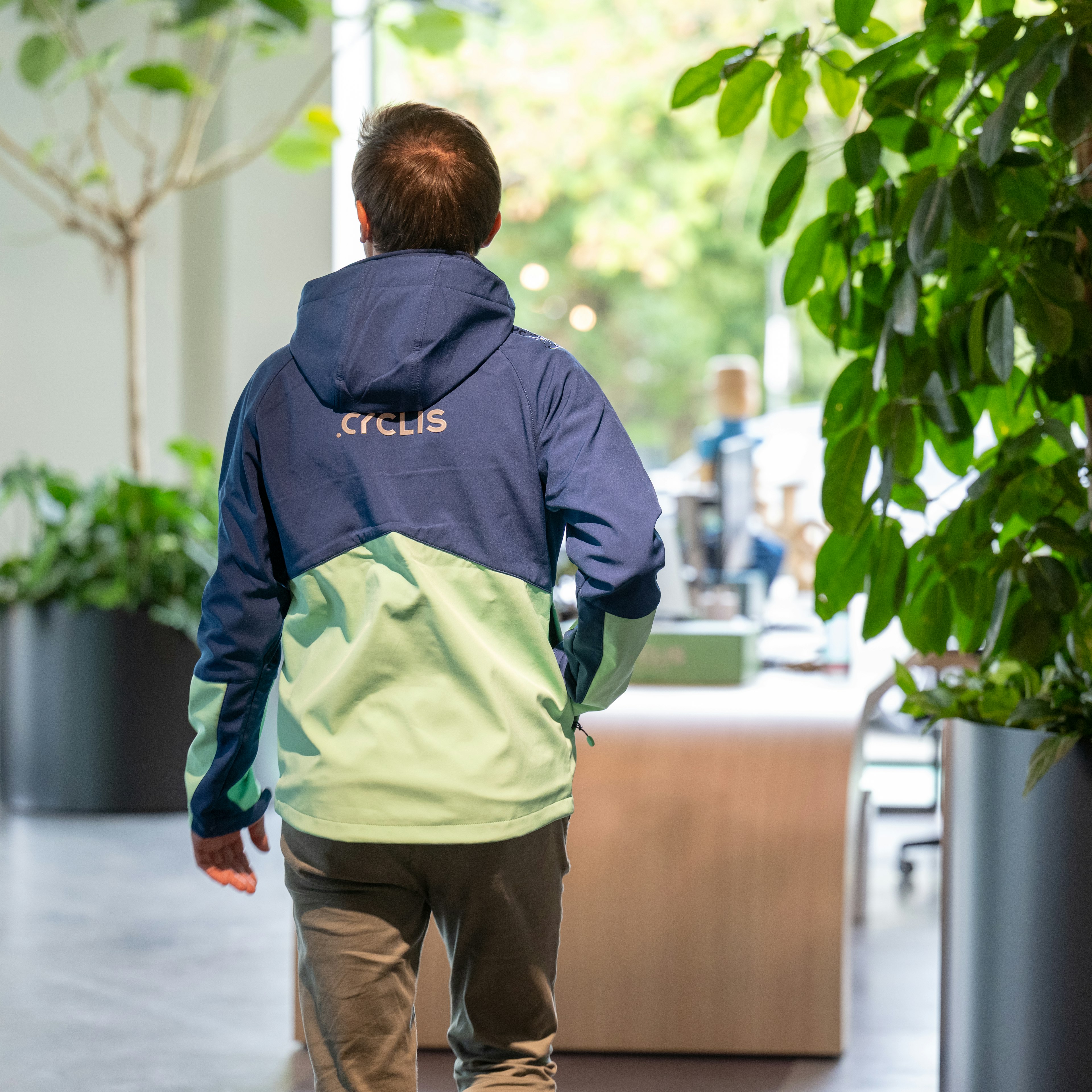 Un employé de Cyclis, vêtu d'une veste aux couleurs de l'entreprise, s'éloigne de la caméra en traversant le bureau baigné de lumière naturelle, en direction de l'espace d'accueil.