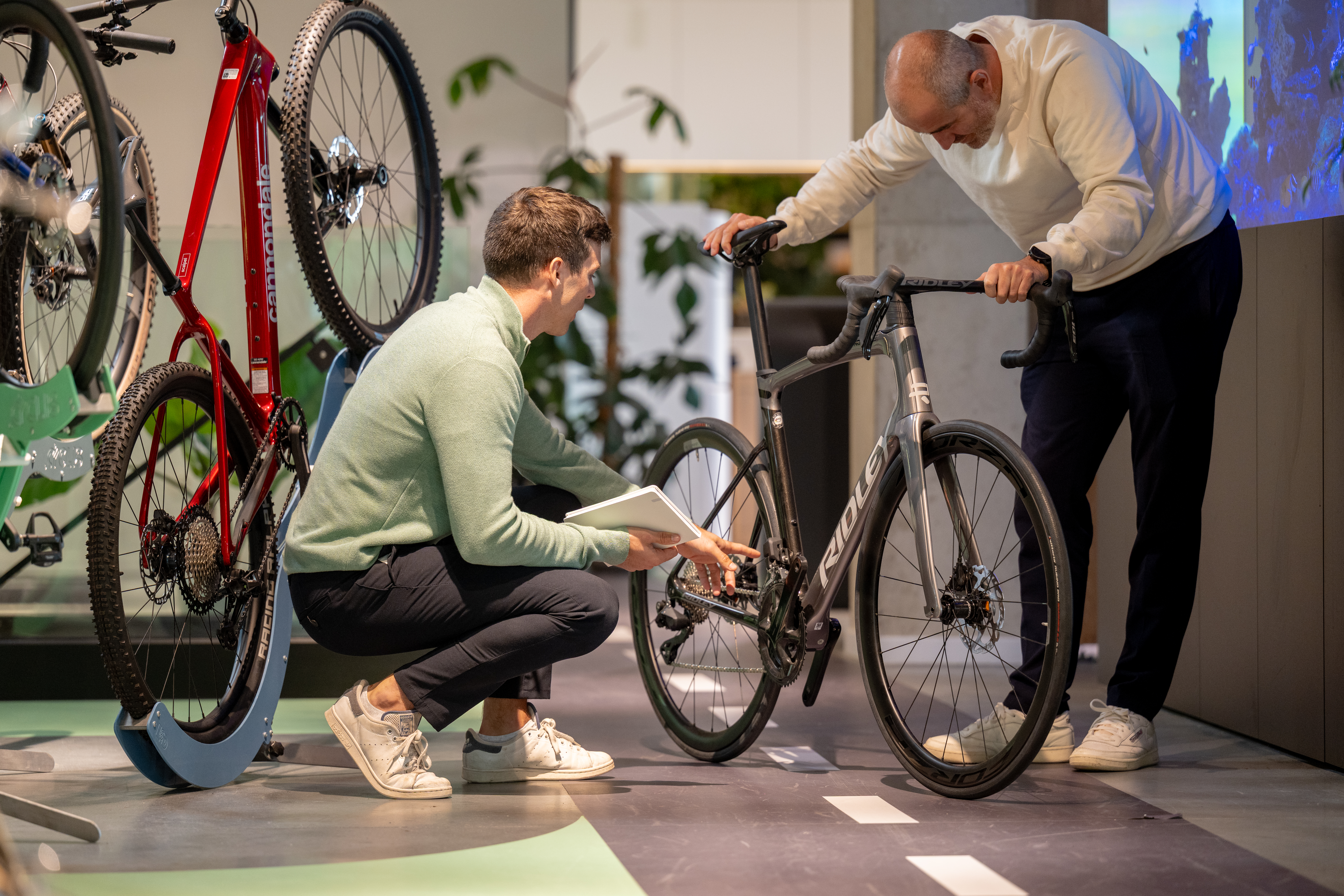 A Cyclis advisor crouches with a tablet to inspect the drivetrain of a Ridley road bike while a customer leans over to look, in the showroom alongside a mounted Cannondale.