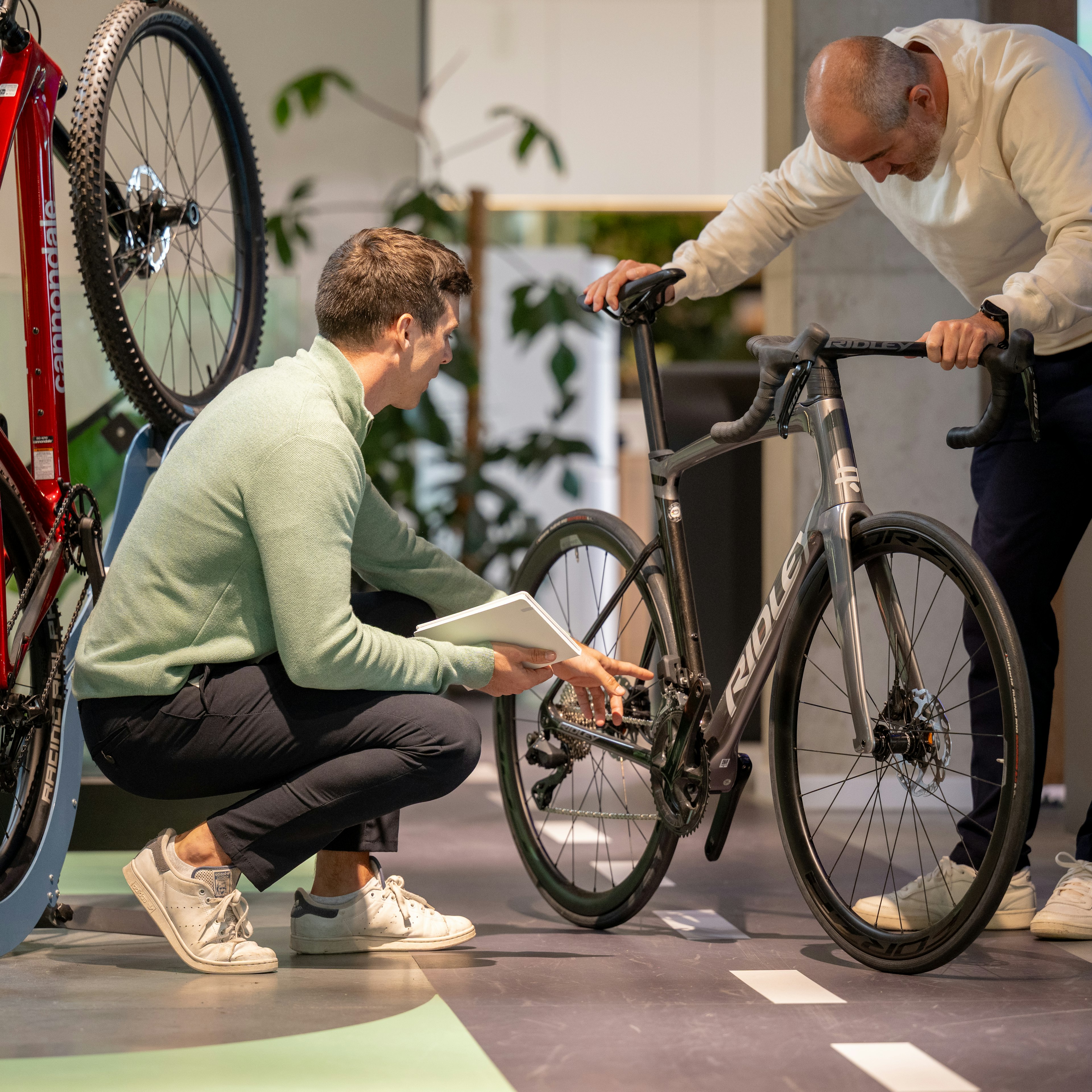 Un conseiller Cyclis est accroupi, une tablette à la main, pour inspecter la transmission d'un vélo de route Ridley, tandis qu'un client se penche pour regarder, dans le showroom, à côté d'un Cannondale exposé.