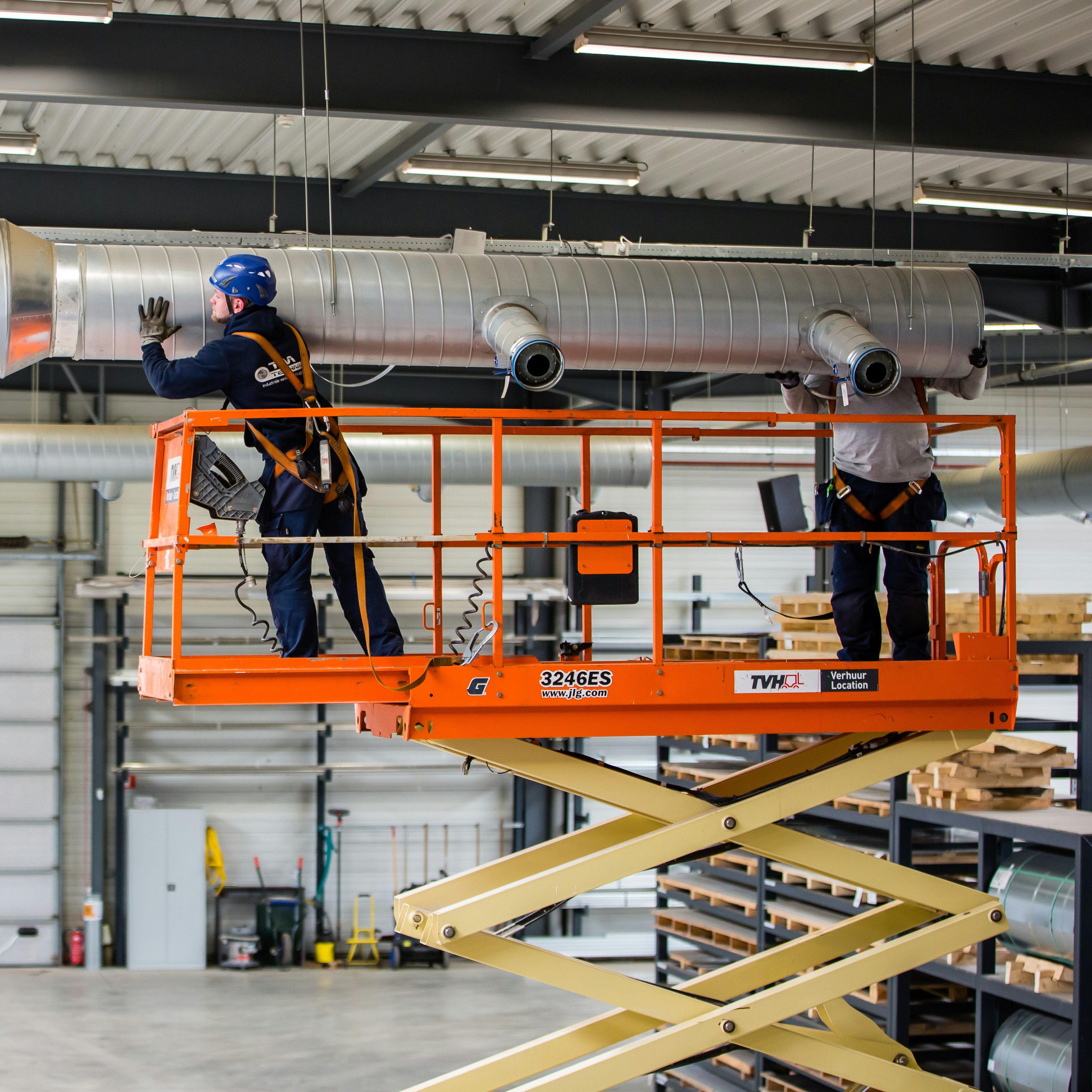 Deux techniciens de TM Technics, installés sur une nacelle à ciseaux orange, posent des conduits spiralés insonorisés au plafond d'un hall industriel.