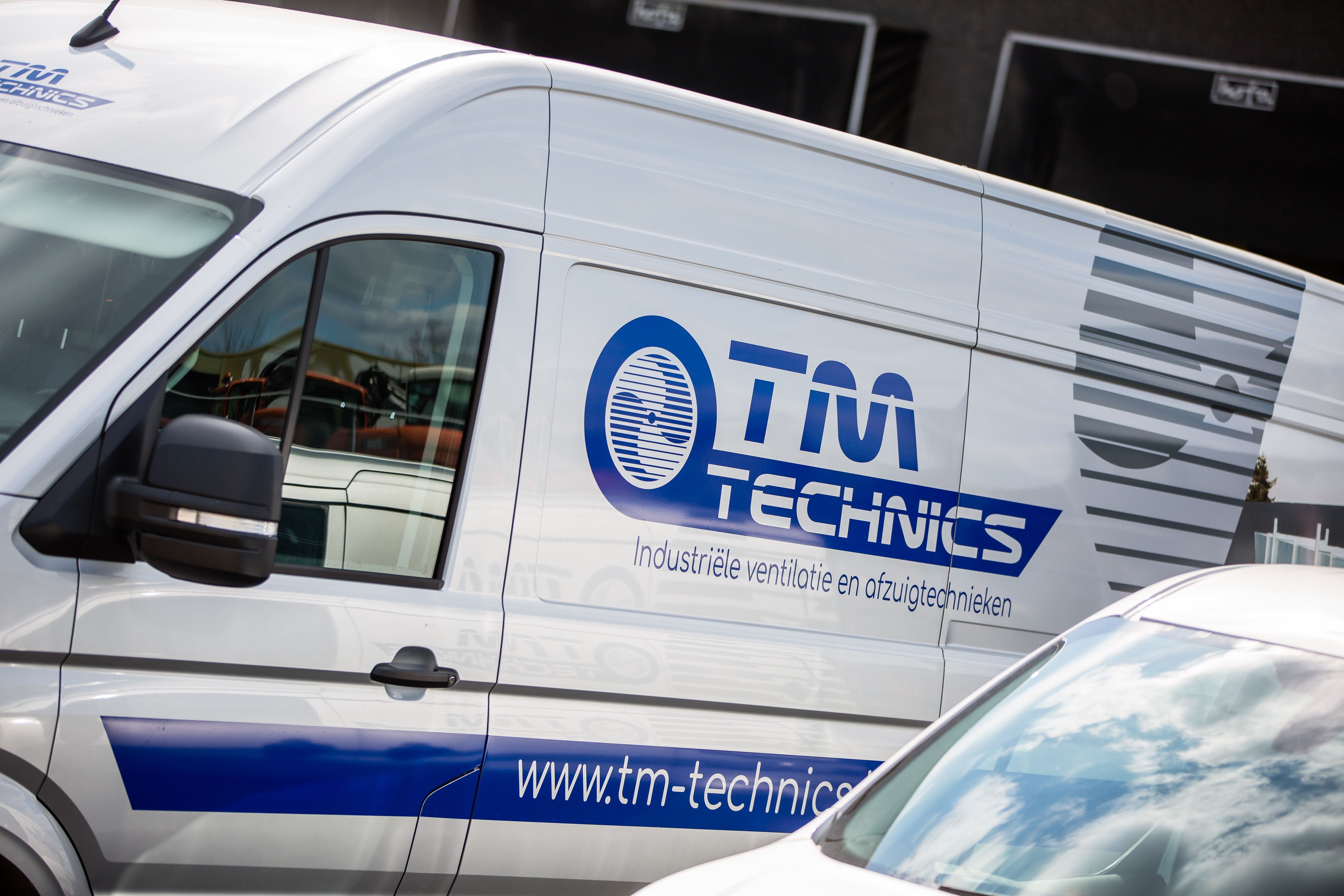 Vue de profil d'une camionnette blanche TM Technics, sur laquelle figurent le logo et le slogan « industriële ventilatie en afzuigtechnieken »