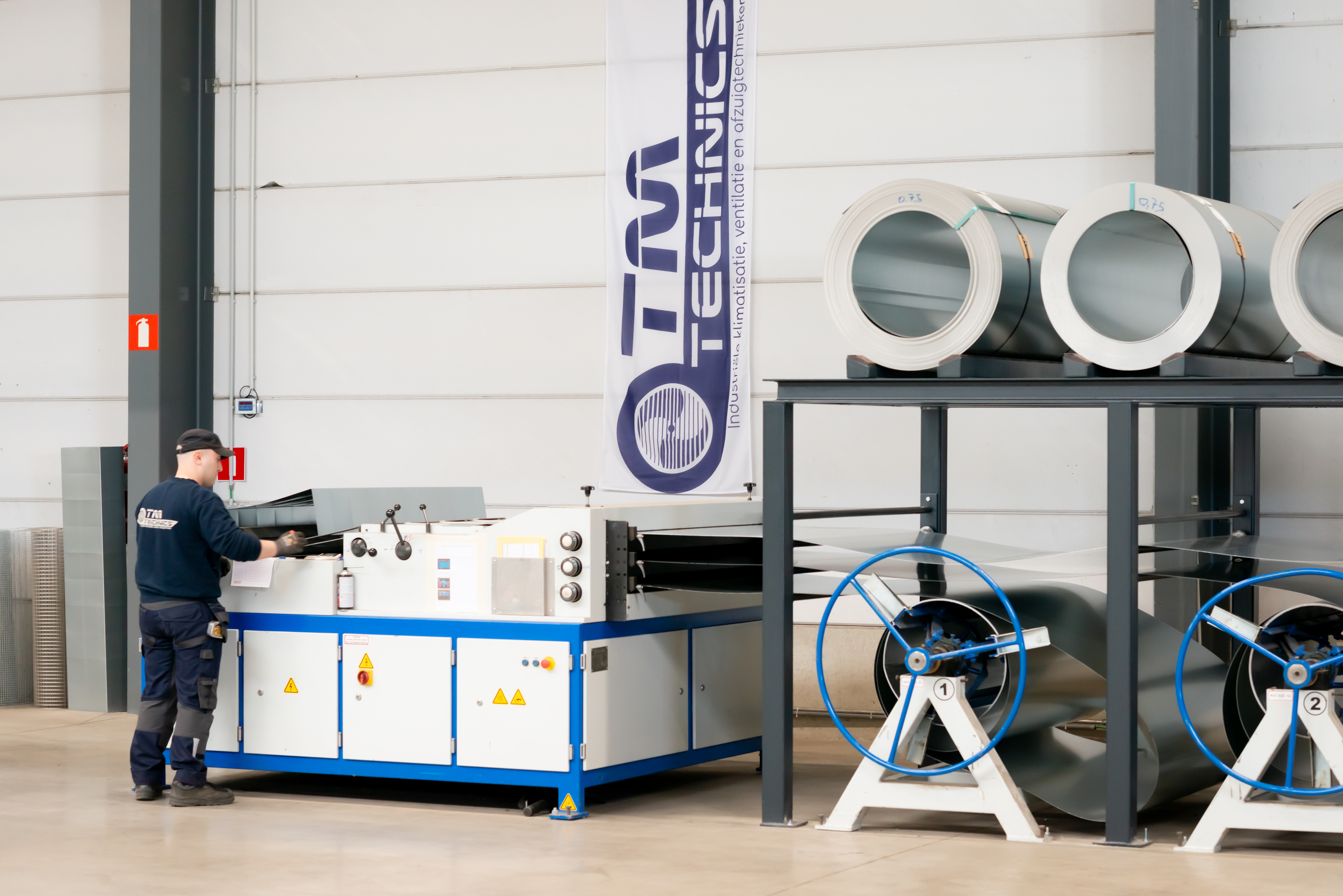 Un technicien de TM Technics travaille sur une ligne de formage de bobines à côté d'une bannière de la marque, avec des bobines d'acier entreposées au-dessus et deux bobines d'alimentation numérotées 1 et 2.