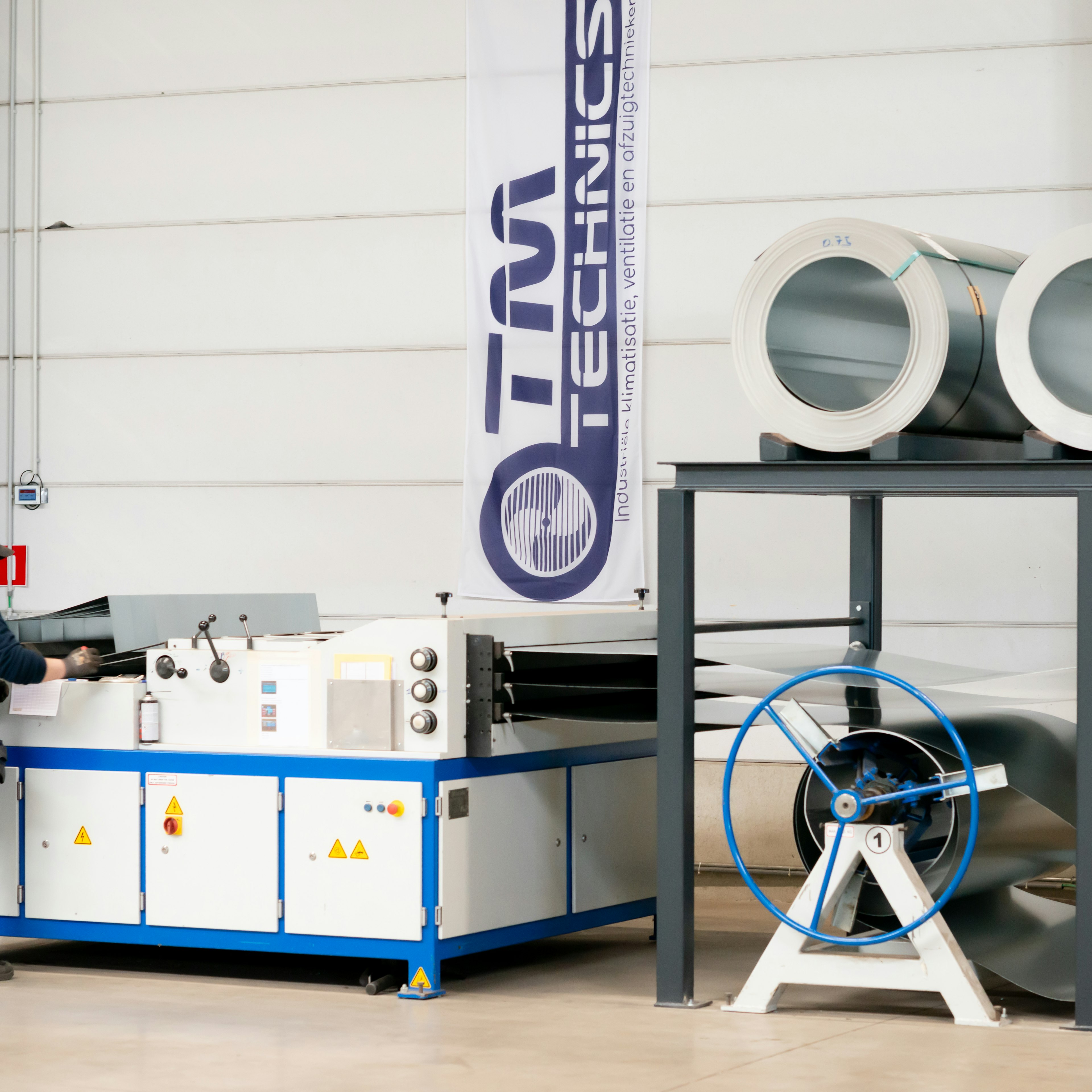 Un technicien de TM Technics travaille sur une ligne de formage de bobines à côté d'une bannière de la marque, avec des bobines d'acier entreposées au-dessus et deux bobines d'alimentation numérotées 1 et 2.