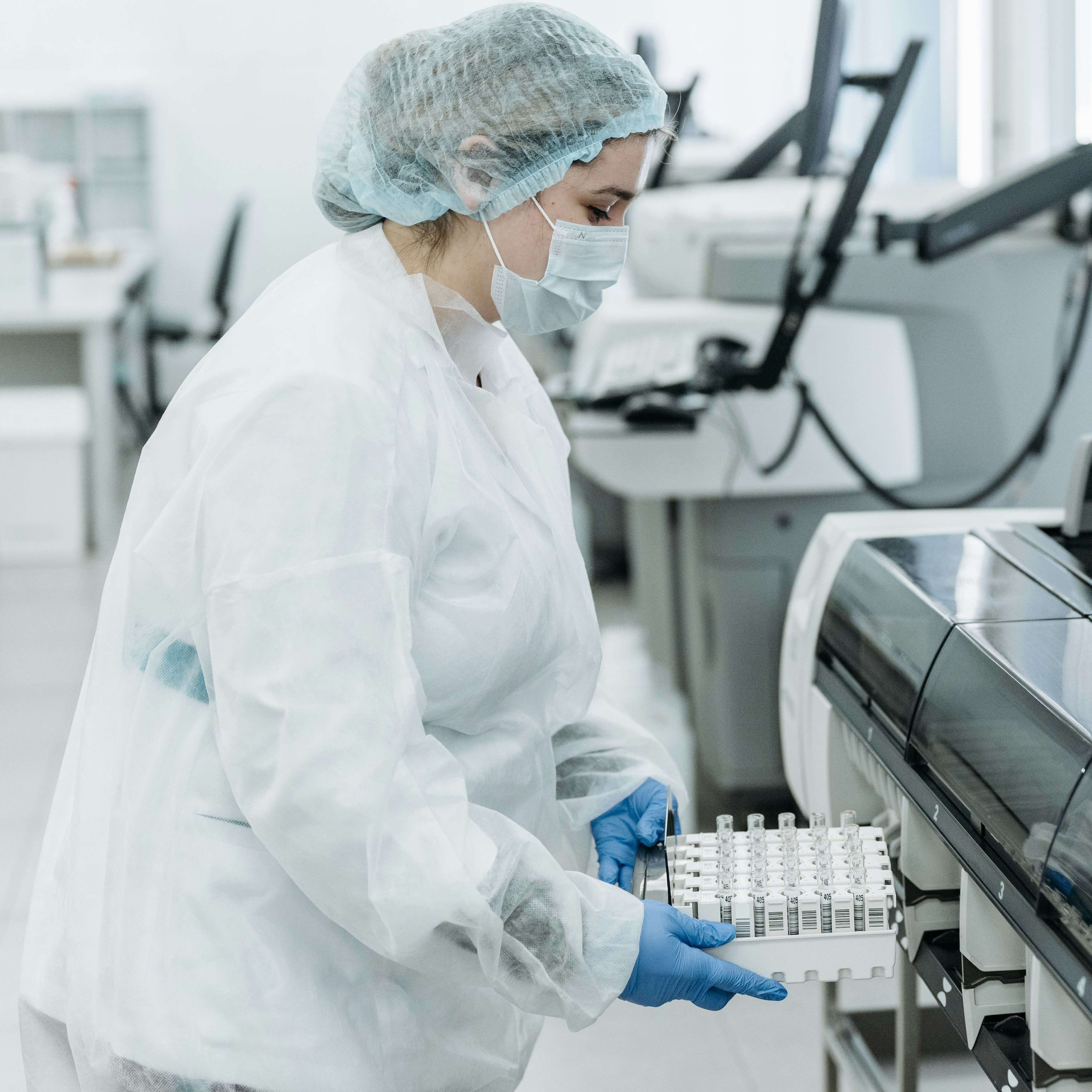 Un technicien de laboratoire, vêtu d'une combinaison blanche et de gants bleus, utilise une pipette pour transférer un liquide bleu dans un portoir de tubes à essai, à côté de flacons contenant des solutions colorées.