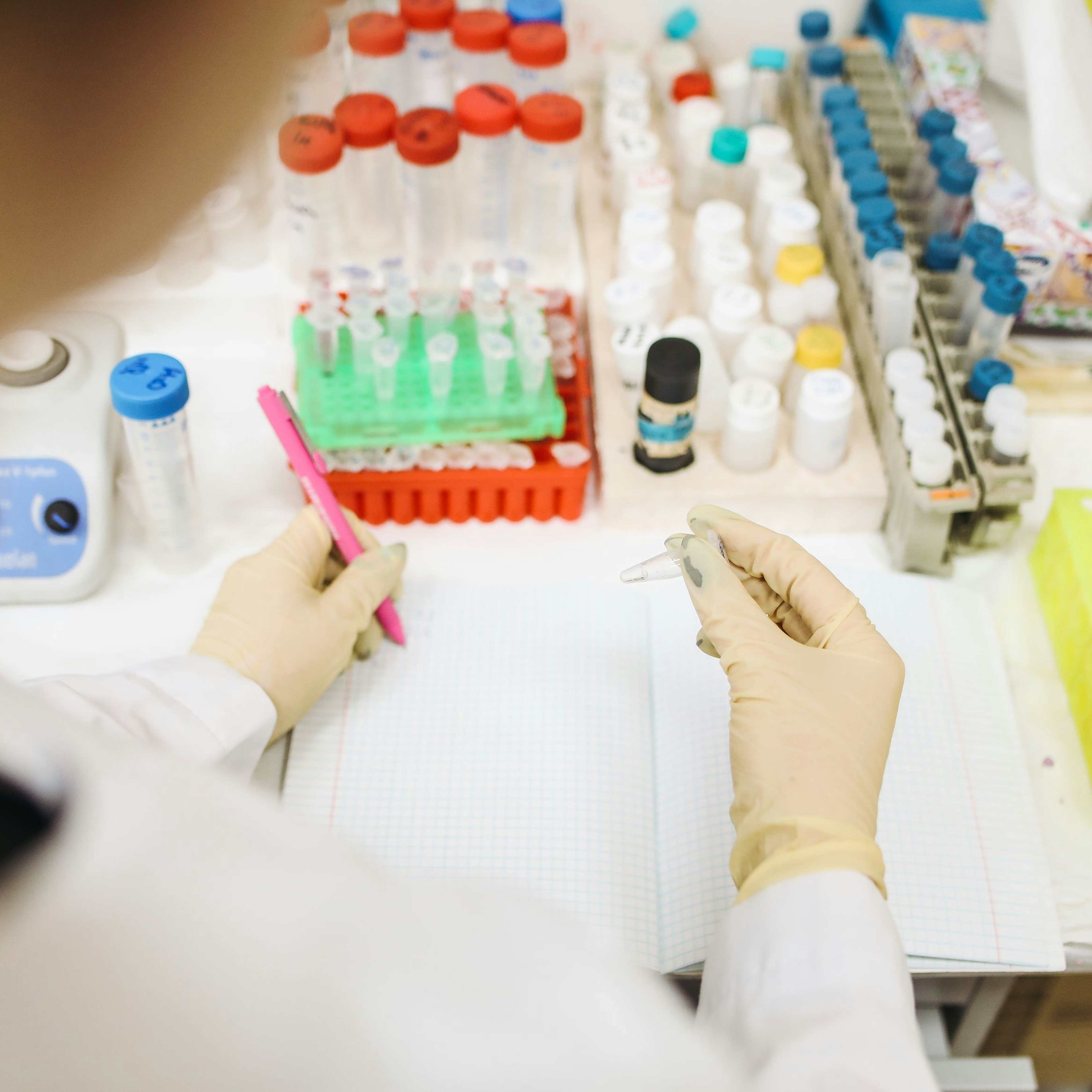 Overhead view of a lab worker in white gloves writing in a notebook while holding a small sample tube, surrounded by racks of capped vials.