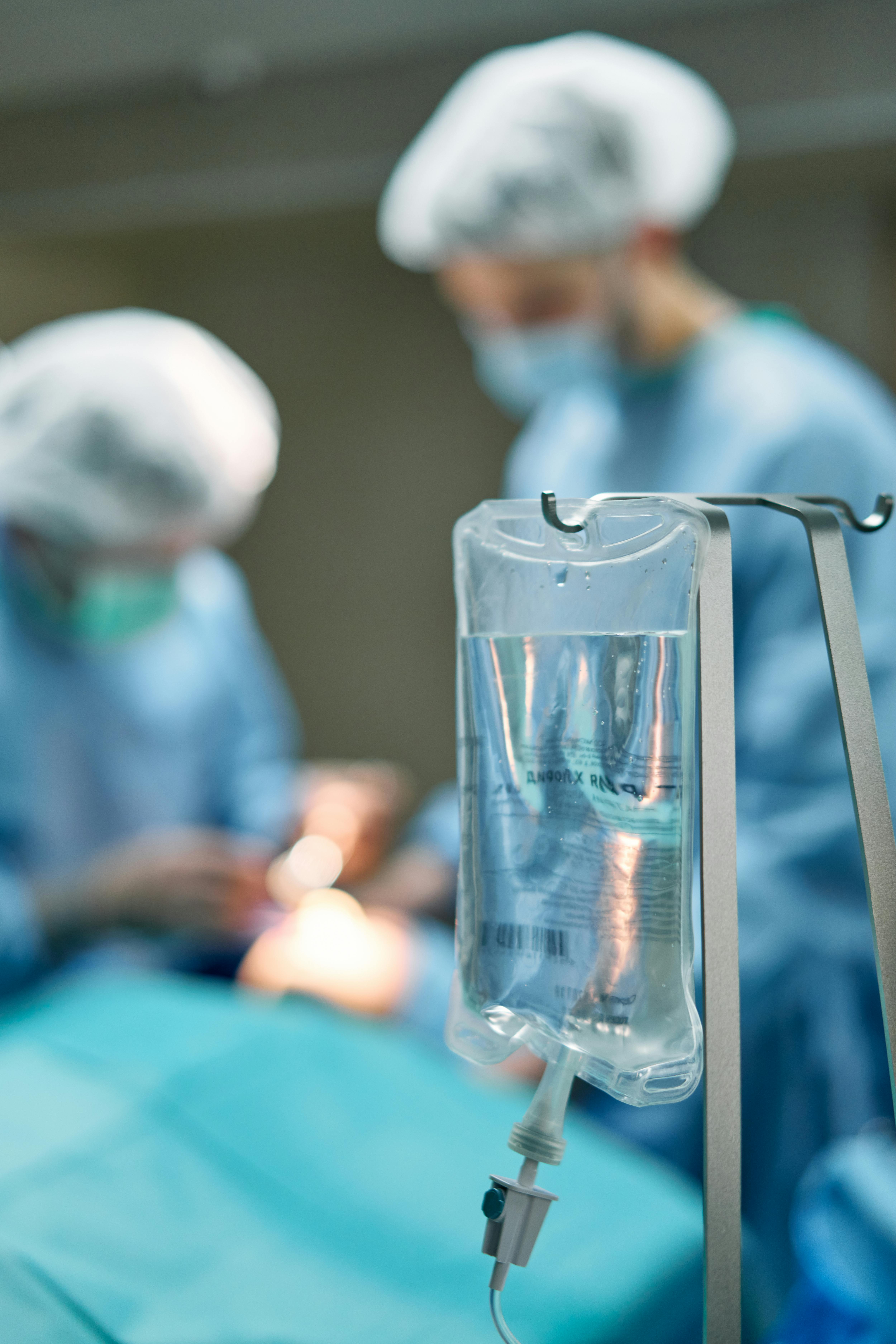 Gros plan sur une poche de perfusion transparente suspendue à une potence métallique au premier plan, avec le tuyau de perfusion et la pince à rouleau visibles en dessous. À l'arrière-plan flou, deux chirurgiens vêtus de blouses bleues, de calots et de masques se penchent sur une table d'opération pendant une intervention. La faible profondeur de champ maintient la mise au point sur la poche de perfusion tandis que l'équipe chirurgicale apparaît légèrement floue. Des tons bleus et turquoise dominent la scène.