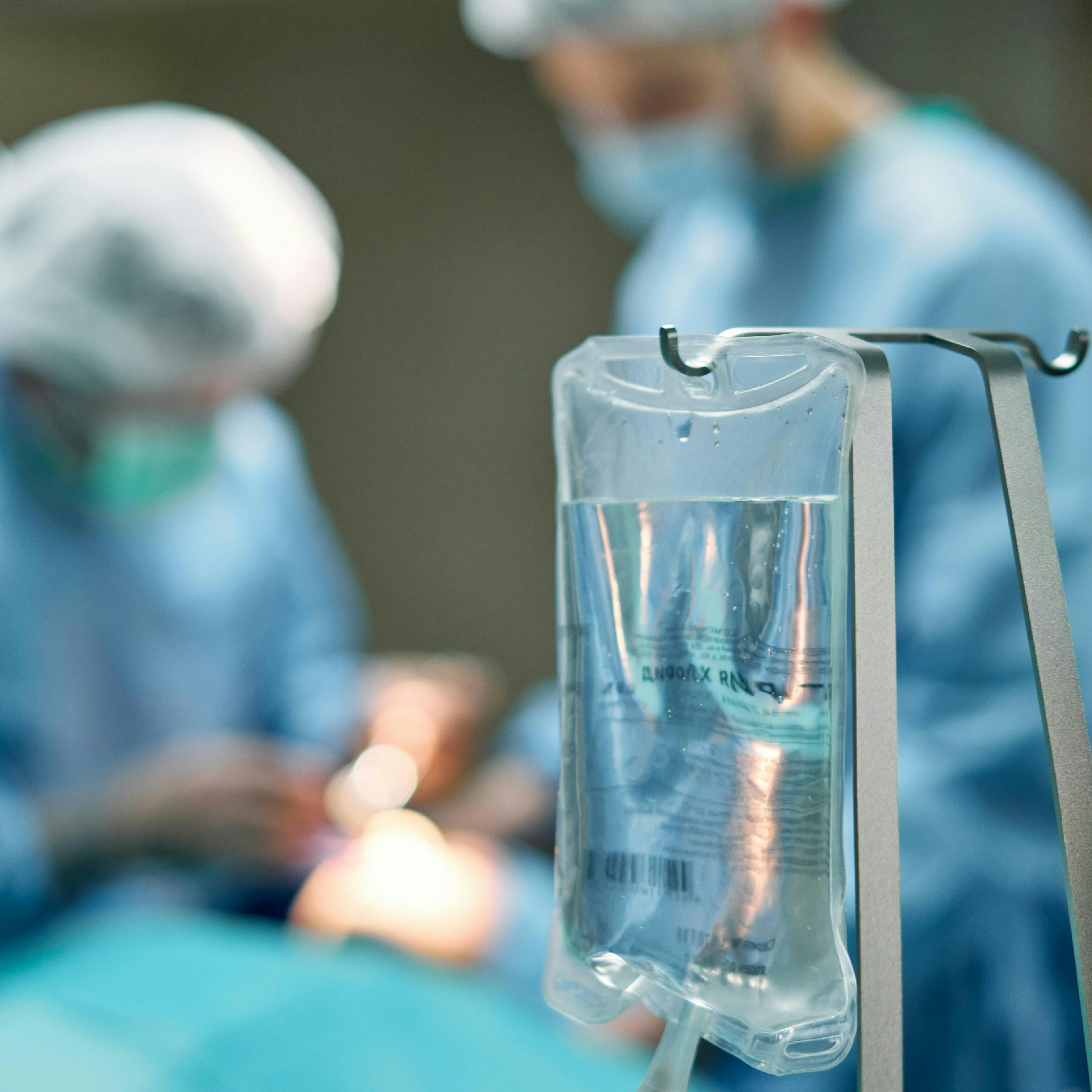 A close-up of a clear IV fluid bag hanging from a metal pole in the sharp foreground, with its drip line and roller clamp visible below. In the blurred background, two surgeons in blue scrubs, caps, and masks lean over an operating table during a procedure. The shallow depth of field keeps the focus on the IV bag while the surgical team works softly out of focus. Cool blue and teal tones dominate the scene.