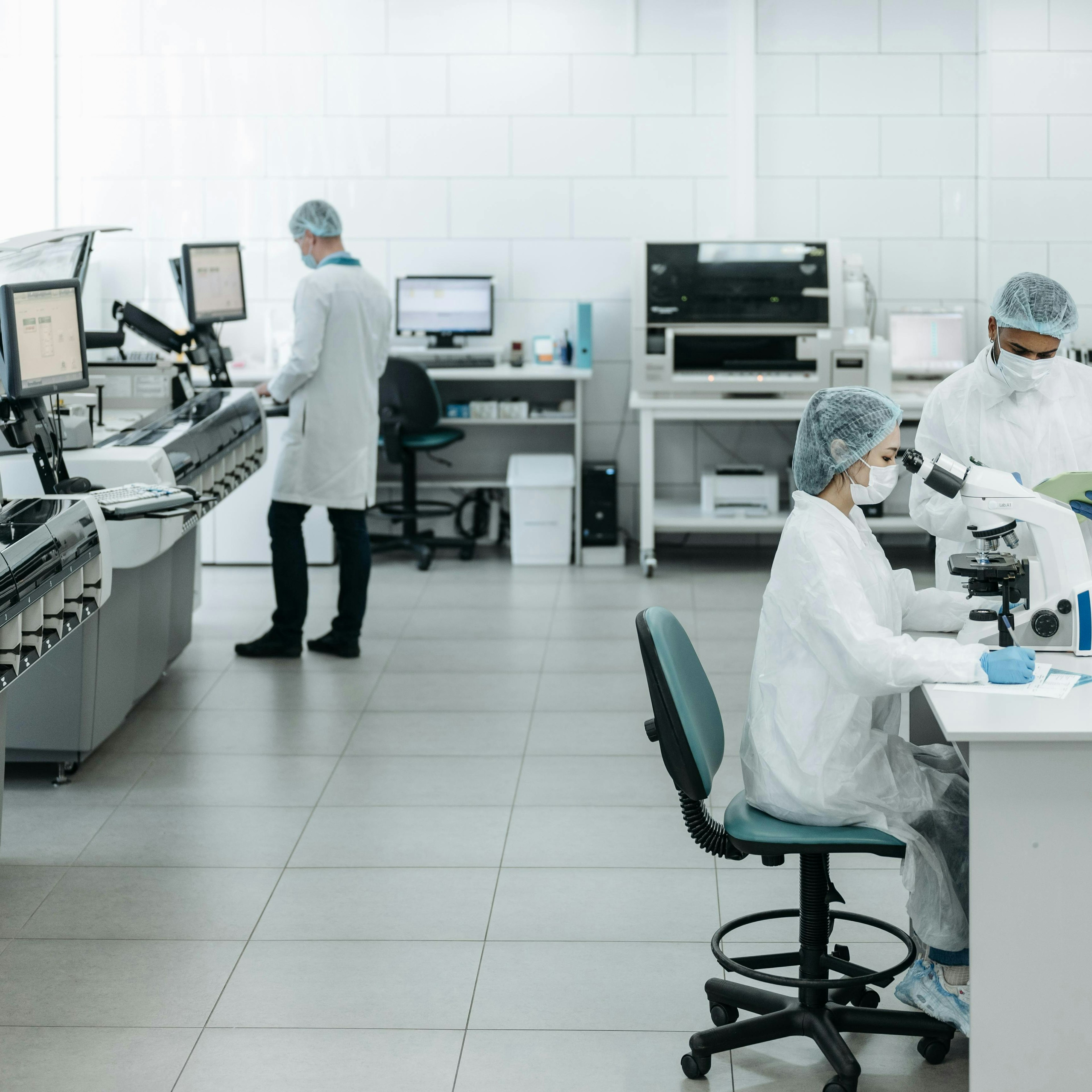 A bright, modern clinical laboratory with white tiled walls and tiled flooring. On the right, a seated technician in a white coat, cap, mask, and blue gloves looks into a microscope, while a colleague standing beside her holds a green folder. To their right, racks of red-capped test tubes sit on the bench. On the left side, a row of large automated analyzers with screens lines the wall, and in the background a technician in a white coat works at a computer station. The overall palette is clean, white, and cool-toned.